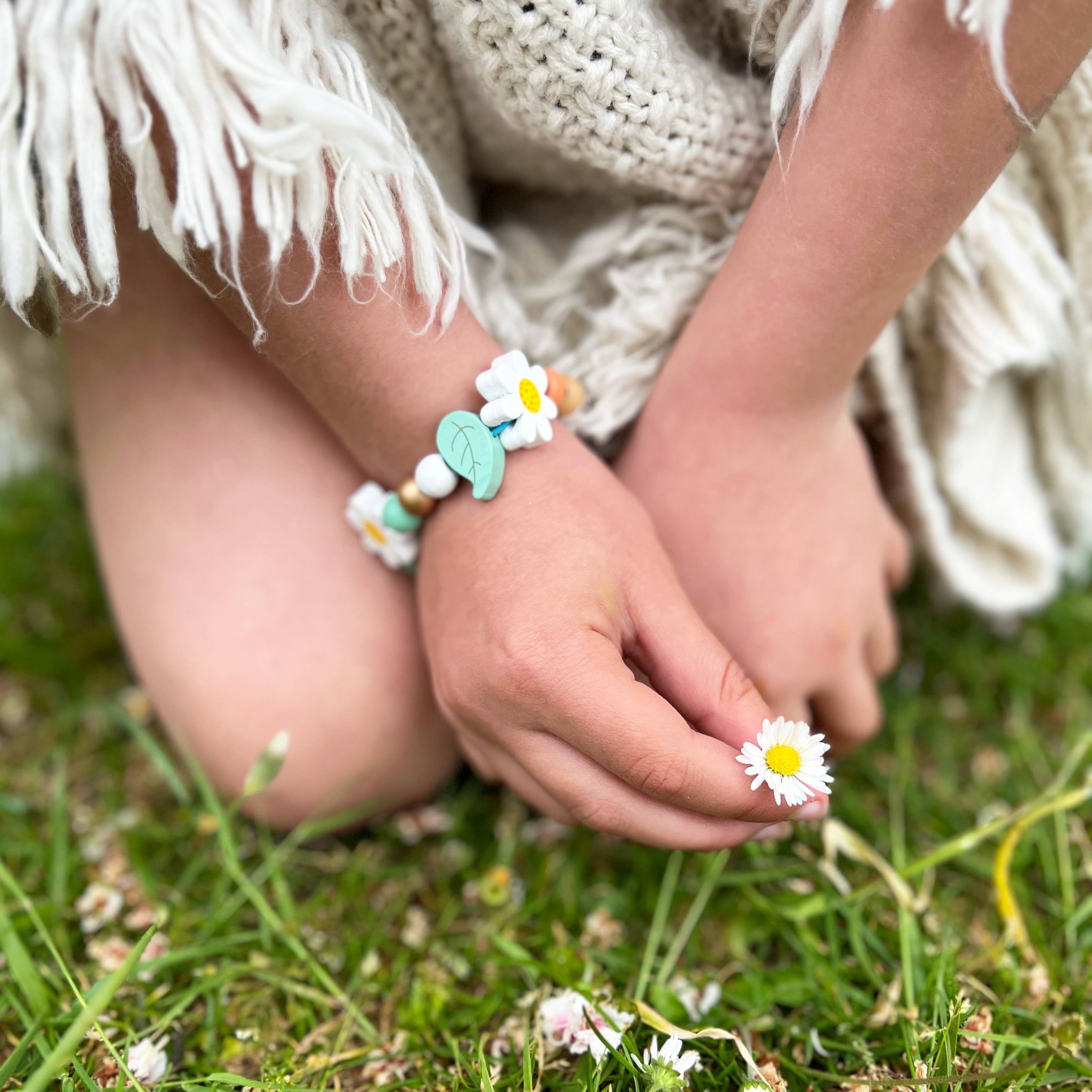Ein Kind kniet im Gras und hält in einer Hand ein kleines weiß-gelbes Gänseblümchen. Es trägt ein Cotton Twist – DIY Armbandset Gänseblümchen, das mit Gänseblümchen und Blättern geschmückt ist. Das Kind ist in ein mit Fransen verziertes, cremefarbenes Gewand gekleidet. Die Szene ruft ein Gefühl von Unschuld und Natur hervor.