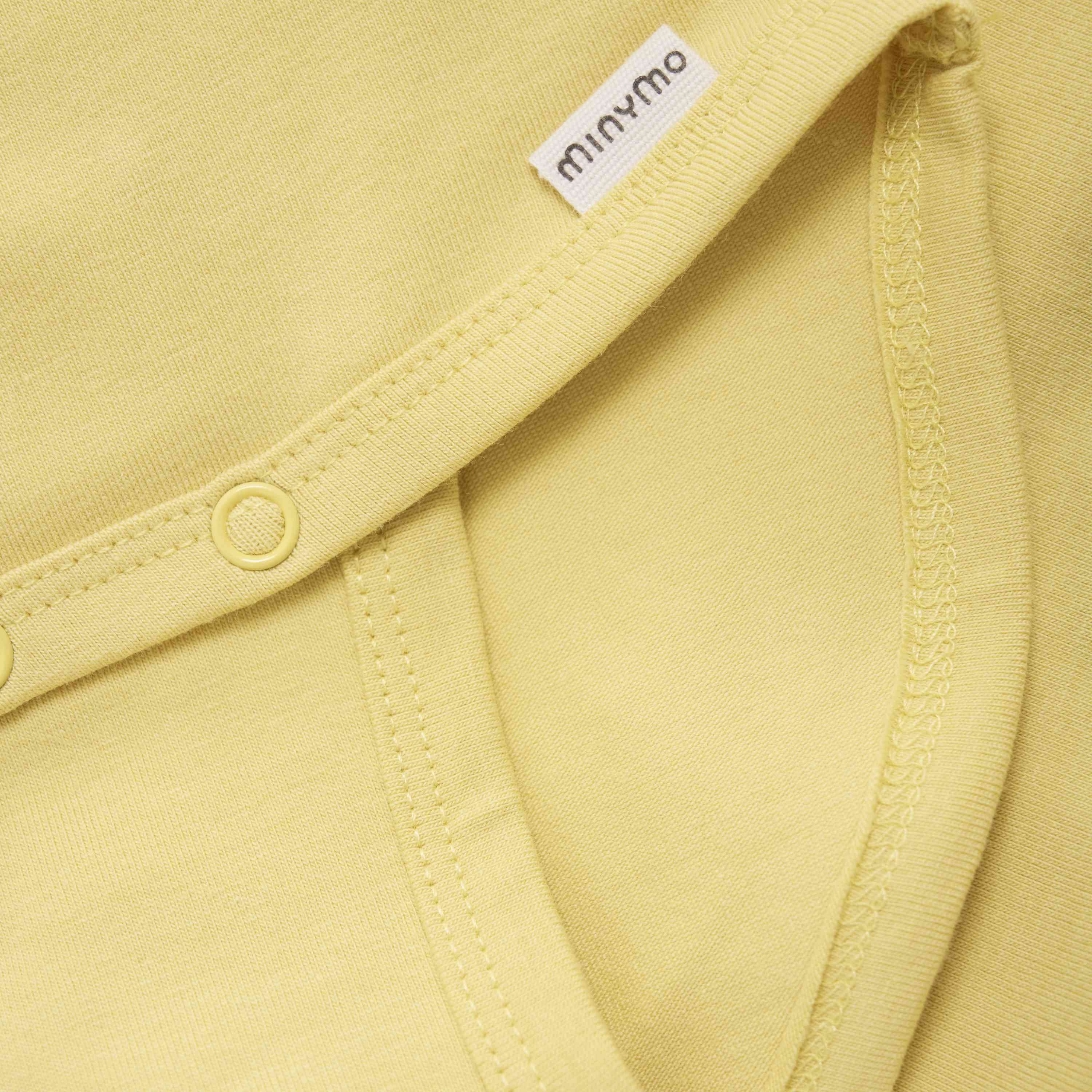 Nahaufnahme von hellgelber Babykleidung mit einem sichtbaren Etikett mit dem Markennamen „Minymo“. Das Kleidungsstück aus weichem GOTS-zertifiziertem Stoff mit sauberen Nähten hat Druckknöpfe entlang der Naht und ist ein praktisches Minymo-Body-Langarm-Design.