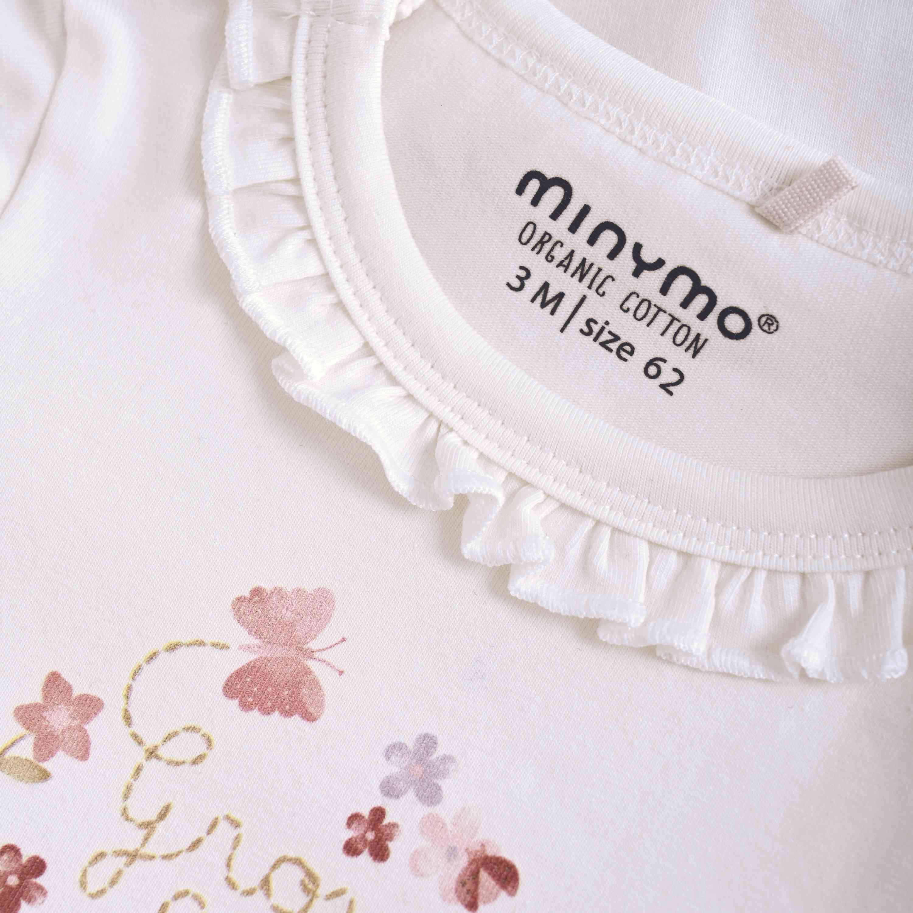 Nahaufnahme des Halsausschnitts und des oberen Brustbereichs eines weißen Babykleidungsstücks aus Bio-Baumwolle mit der Aufschrift „minymo“ und der Größe 3M/62. Dieser Minymo-Body langarm hat einen Rüschenrand entlang des Halsausschnitts mit pastellfarbenen Blumen- und Schmetterlingsdrucken, begleitet vom Text „Growing...“. Ideal für alle, die nachhaltige Babybodys suchen.