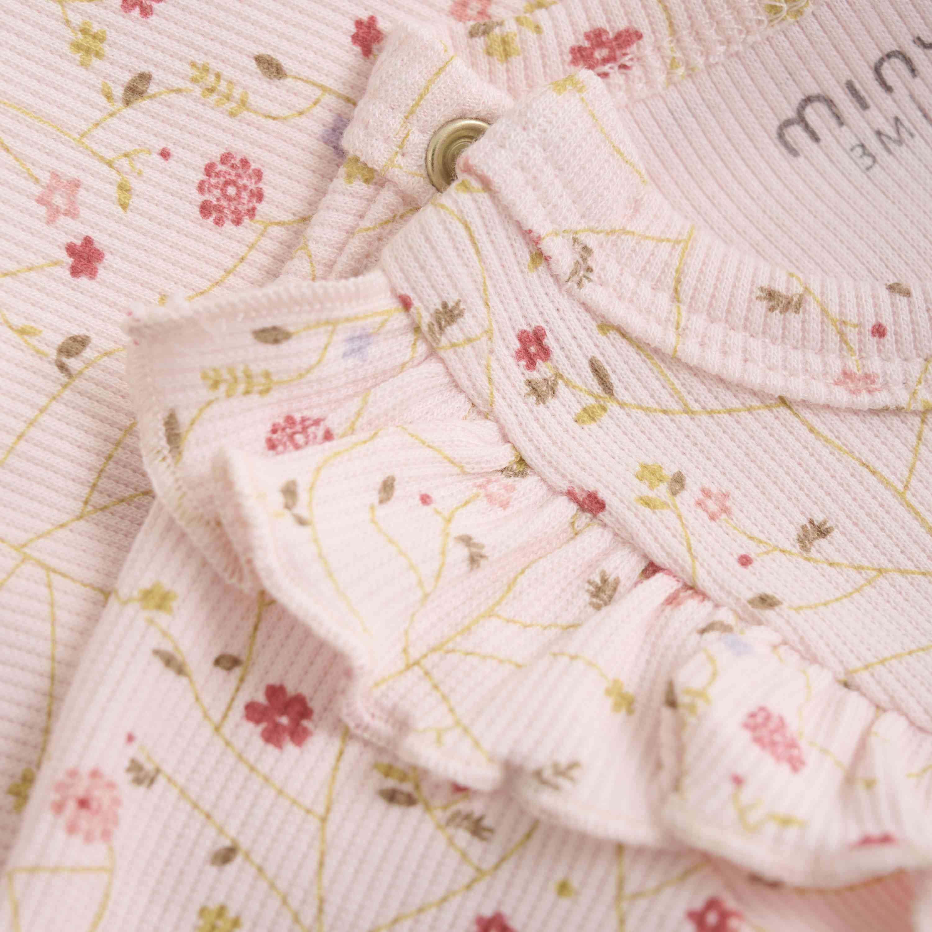 Nahaufnahme eines Minymo Minymo - Body kurzarm aus Rippstoff, einem hellrosa Baby-Strampler mit Blumenmuster aus kleinen rosa und gelben Blumen. Der GOTS-zertifizierte Stoff ist gerippt und hat Rüschendetails in der Nähe des Ausschnitts, wobei ein Druckknopf teilweise sichtbar ist.