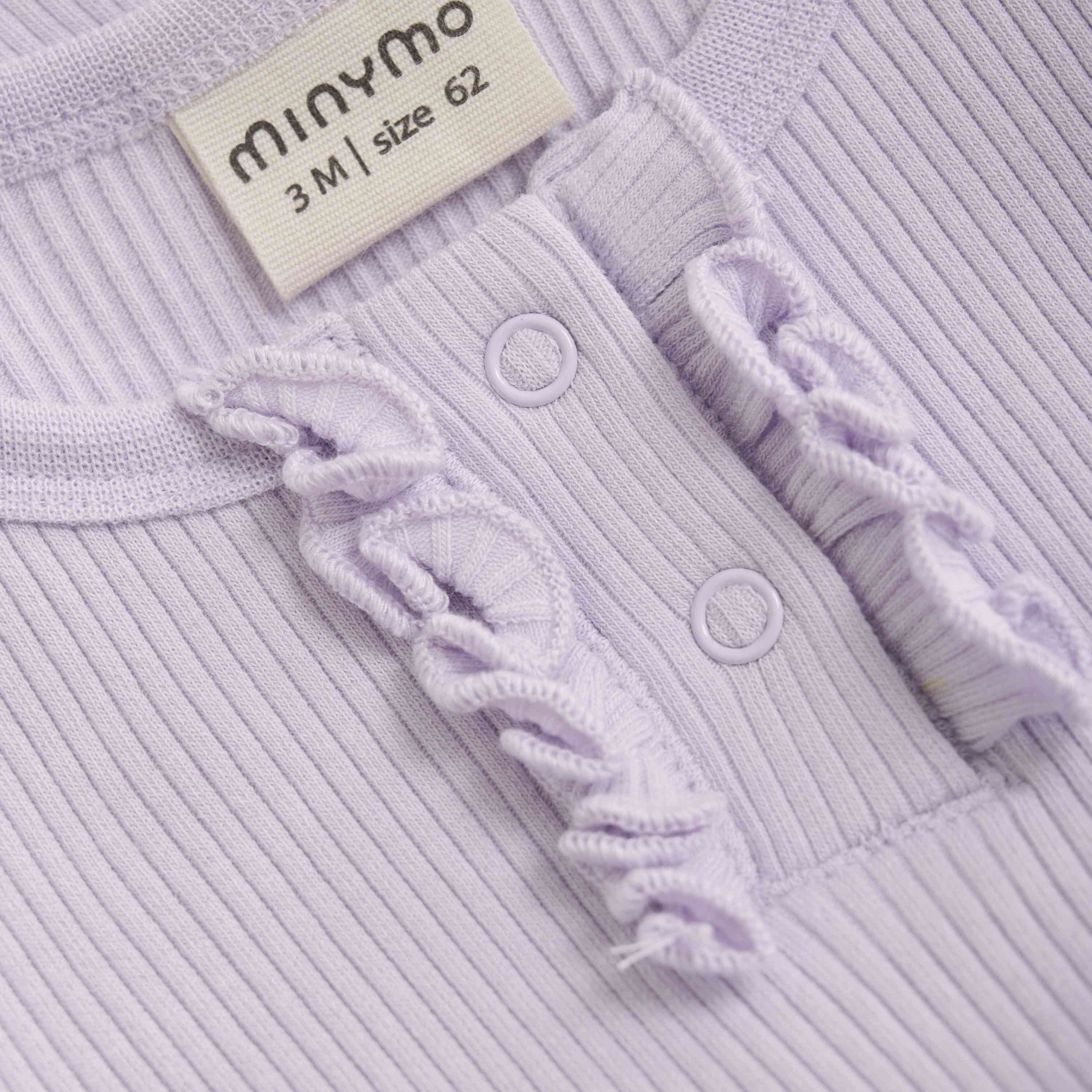 Eine Nahaufnahme eines hellvioletten gerippten Babykleidungsstücks von Minymo. Am Halsausschnitt befindet sich ein Etikett mit der Aufschrift „minymo 3M | Größe 62“. Das Kleidungsstück verfügt über zwei kleine Druckknöpfe und Rüschendetails entlang der Knopfleiste, perfekt für alle, die einen stilvollen Minymo - Body kurzarm aus Rippstoff suchen.