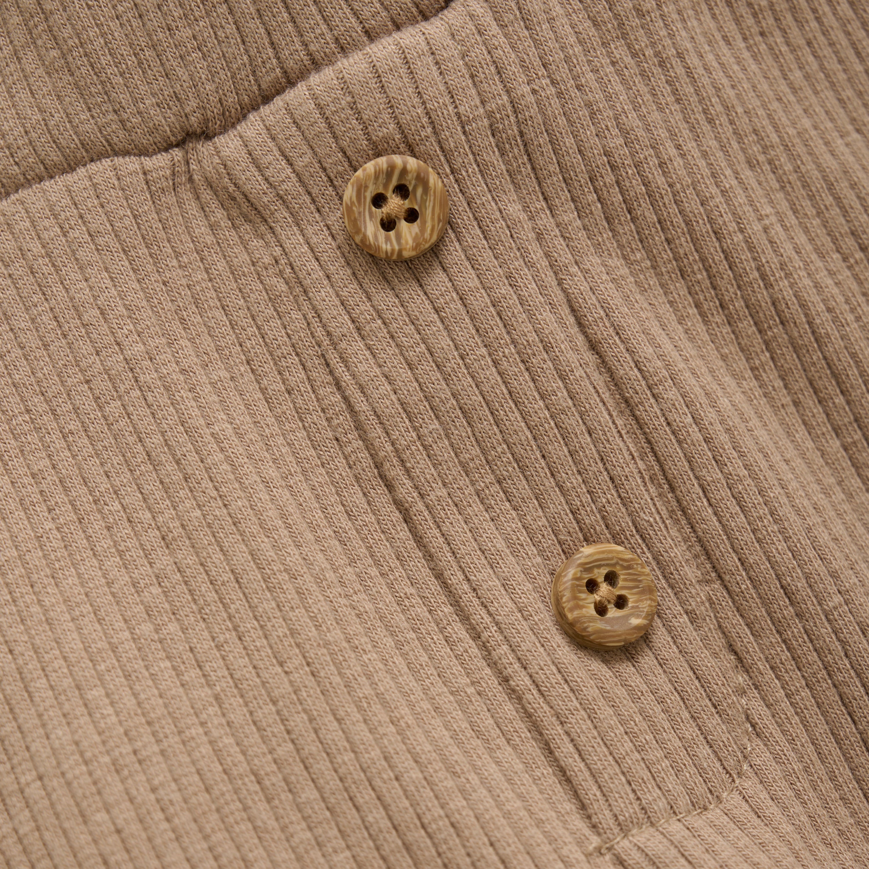 Nahaufnahme eines beigen Rippstoffs mit zwei braunen Knöpfen mit jeweils vier Löchern, die auf den Stoff genäht sind und seine Struktur und Details hervorheben. Der weiche Stoff aus TENCEL™ Modal-Fasern passt perfekt zu den gemütlichen Minymo - Kurze Hosen aus Ripp-Stoff mit einem elastischen Bund für zusätzlichen Komfort von Minymo.
