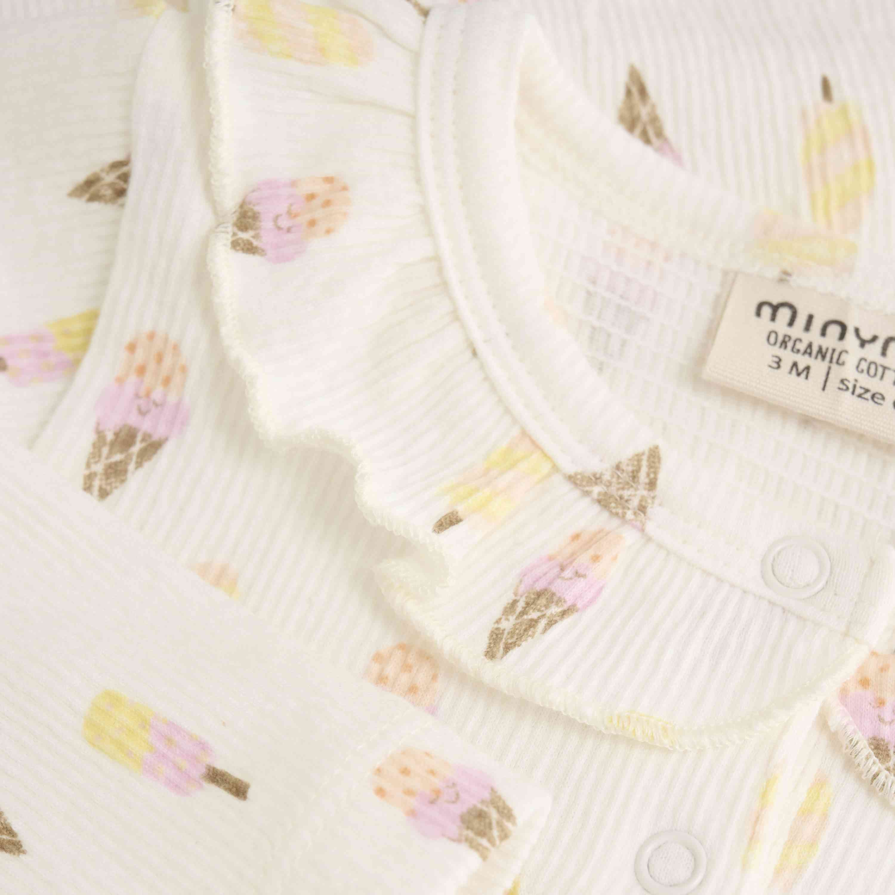 Nahaufnahme eines weißen Minymo - Langarmbodys mit Allover-Print mit zarter Waffelstruktur. Der Body kurzarm ist mit kleinen, bunten Eistüten-Prints verziert. Er verfügt über einen gerüschten Ausschnitt und ein Etikett mit der Aufschrift „Minymo“ aus Bio-Baumwolle, Größe 3M, GOTS-zertifiziert.