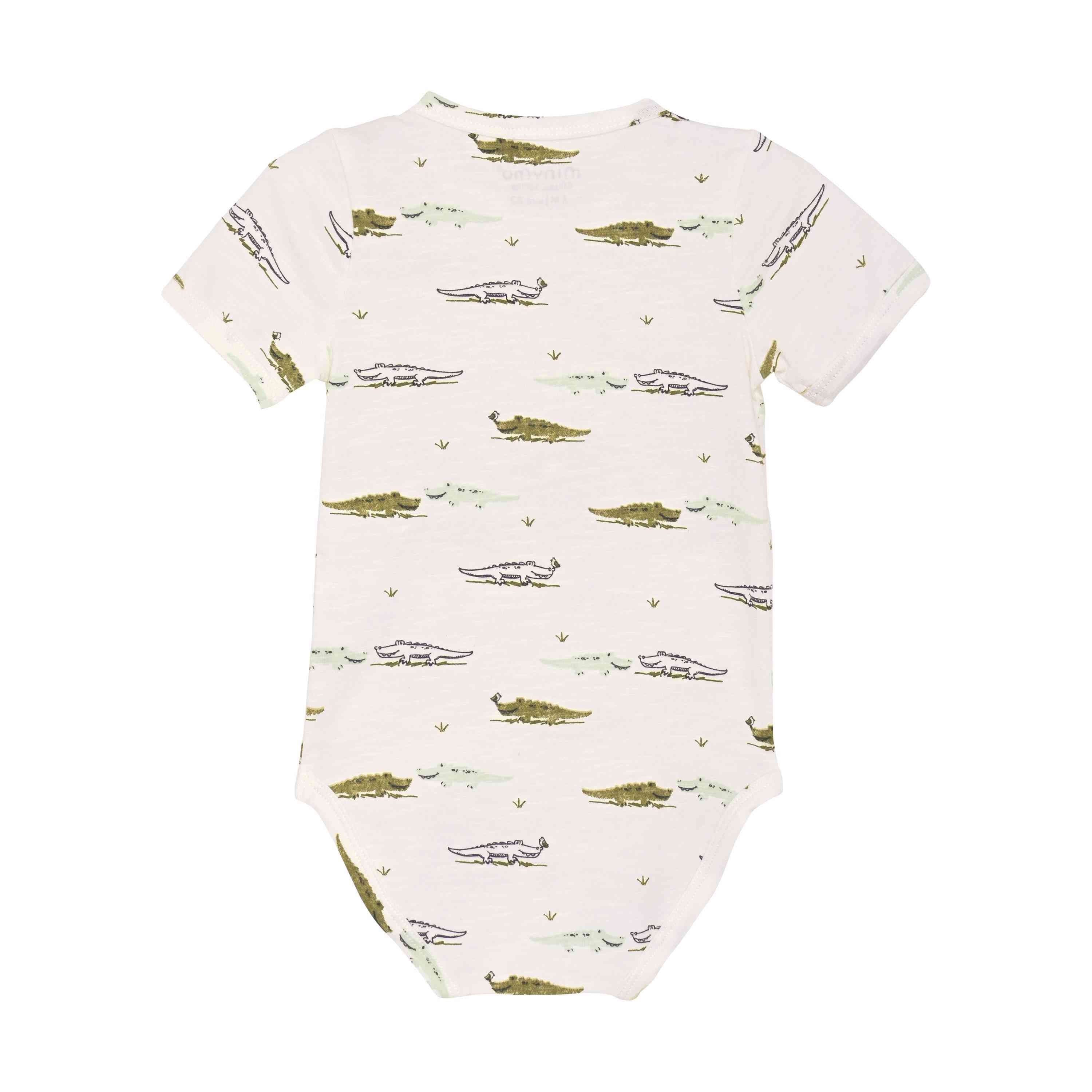 Ein weißer Minymo - Body kurzarm mit Allover-Print-Strampler mit einem Muster aus grünen Krokodilen in verschiedenen Positionen. Die Krokodile sind gleichmäßig über den Bambusstoff verteilt und sorgen für einen Hauch von Minimalismus. Auf dem Bild ist die Rückseite des Stramplers zu sehen.