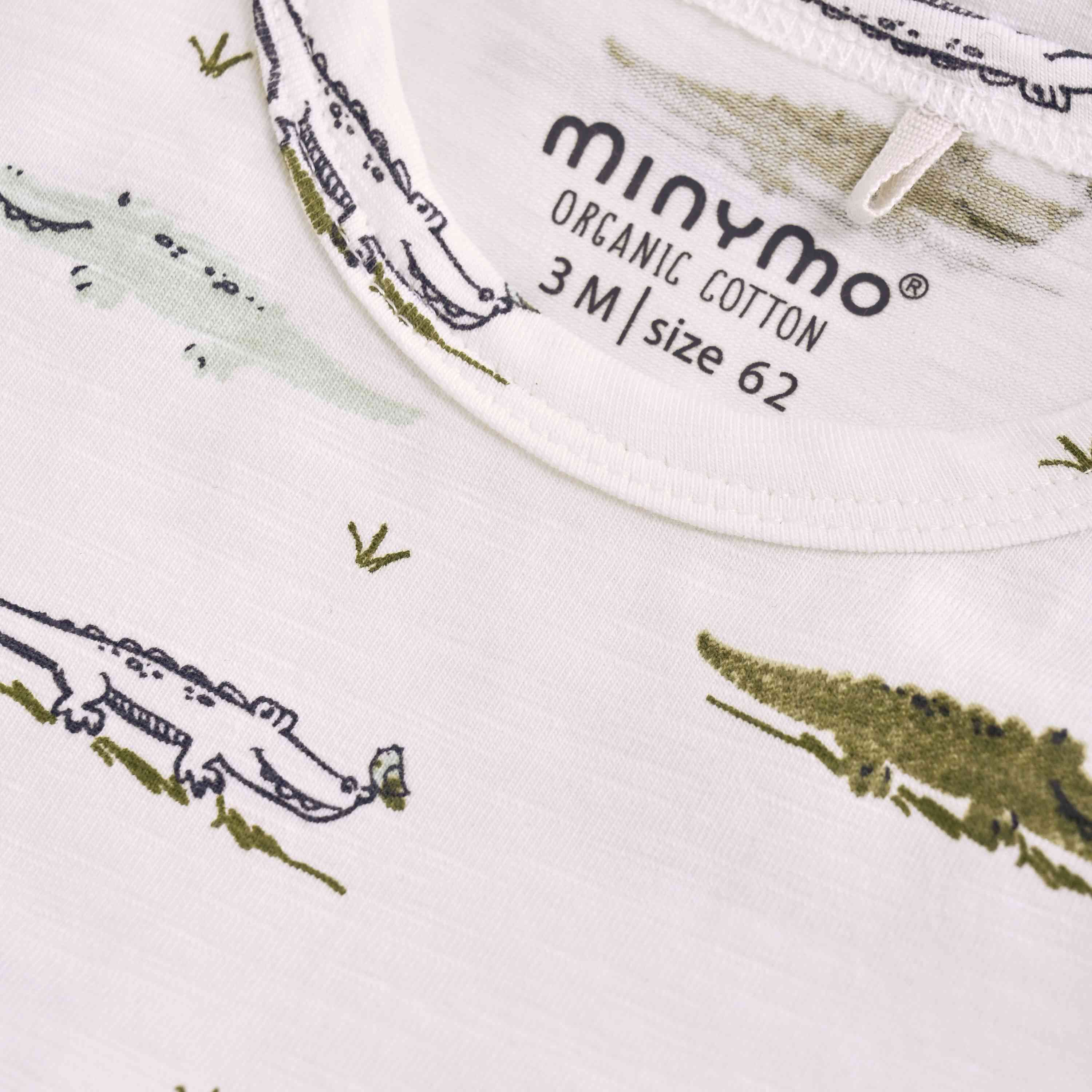 Nahaufnahme eines Babybodys aus weißem Stoff mit grünem Krokodilmuster. Auf dem Etikett steht in schwarzer Schrift „Minymo - Body kurzarm mit Allover-Print 3M Größe 62“. Der Stoff wirkt weich und gut verarbeitet, passend für ein Baby.