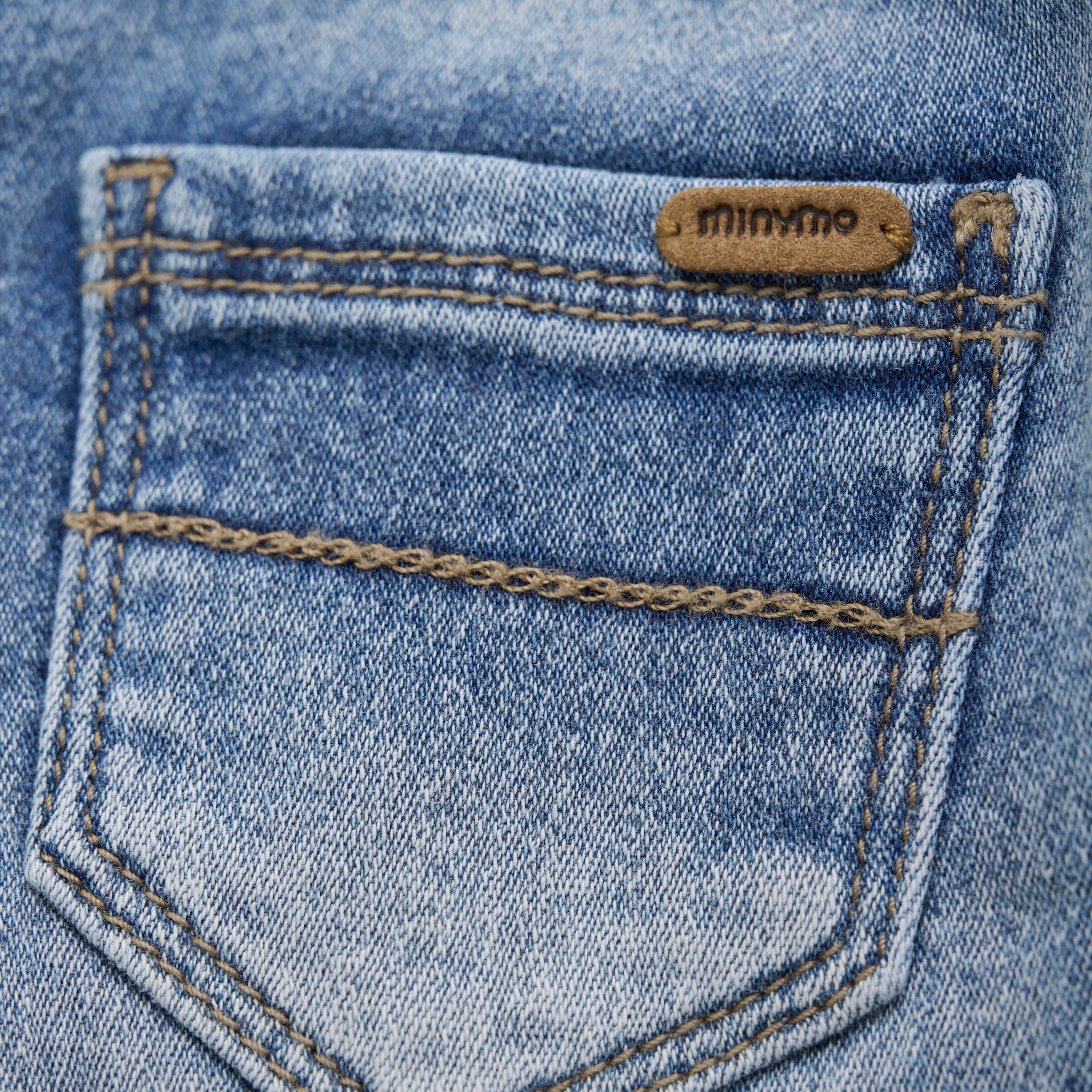 Nahaufnahme einer Gesäßtasche aus blauem Denim mit sichtbaren Nähten. Die Tasche ist mit einem kleinen braunen Lederaufnäher versehen, auf dem der Markenname „Minymo“ eingeprägt ist. Das Denimmaterial hat eine leicht verblichene, abgenutzte Textur, die an die bequemen Minymo - Kurze Hosen erinnert.