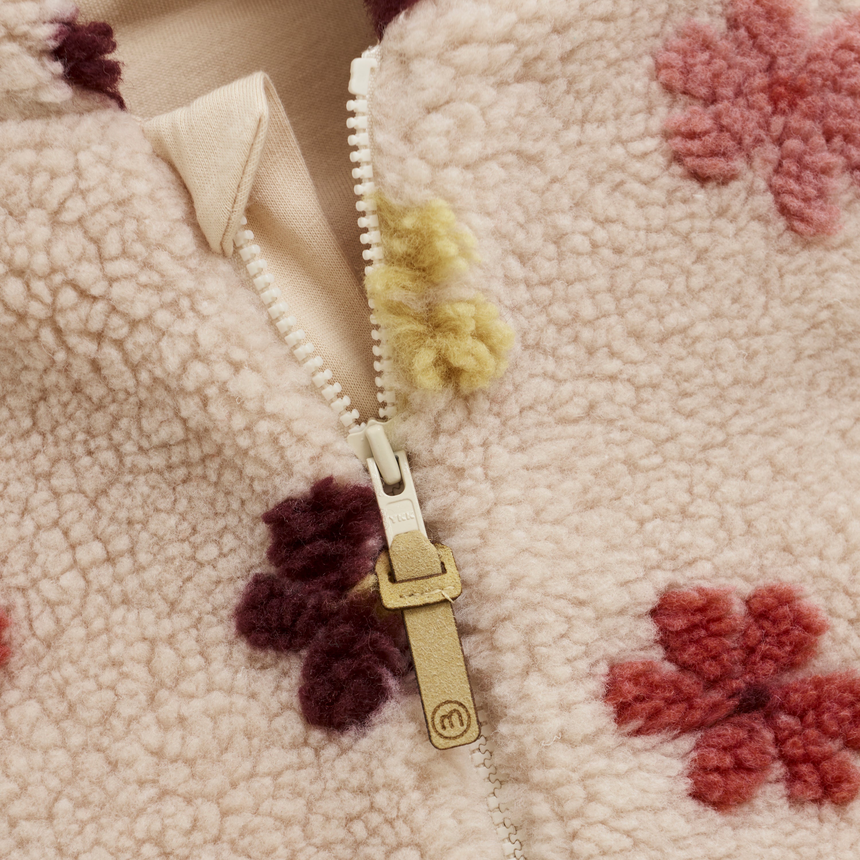 Nahaufnahme des flauschigen Stoffes der Minymo-Jacke, in deren Mitte sich ein Reißverschluss mit einer Lederlasche befindet. Das beigefarbene Kleidungsstück ist mit Blumenmustern in Rot-, Rosa-, Lila- und Gelbtönen verziert. Der teilweise geöffnete Reißverschluss gibt den Blick auf eine darunterliegende beige Schicht frei.