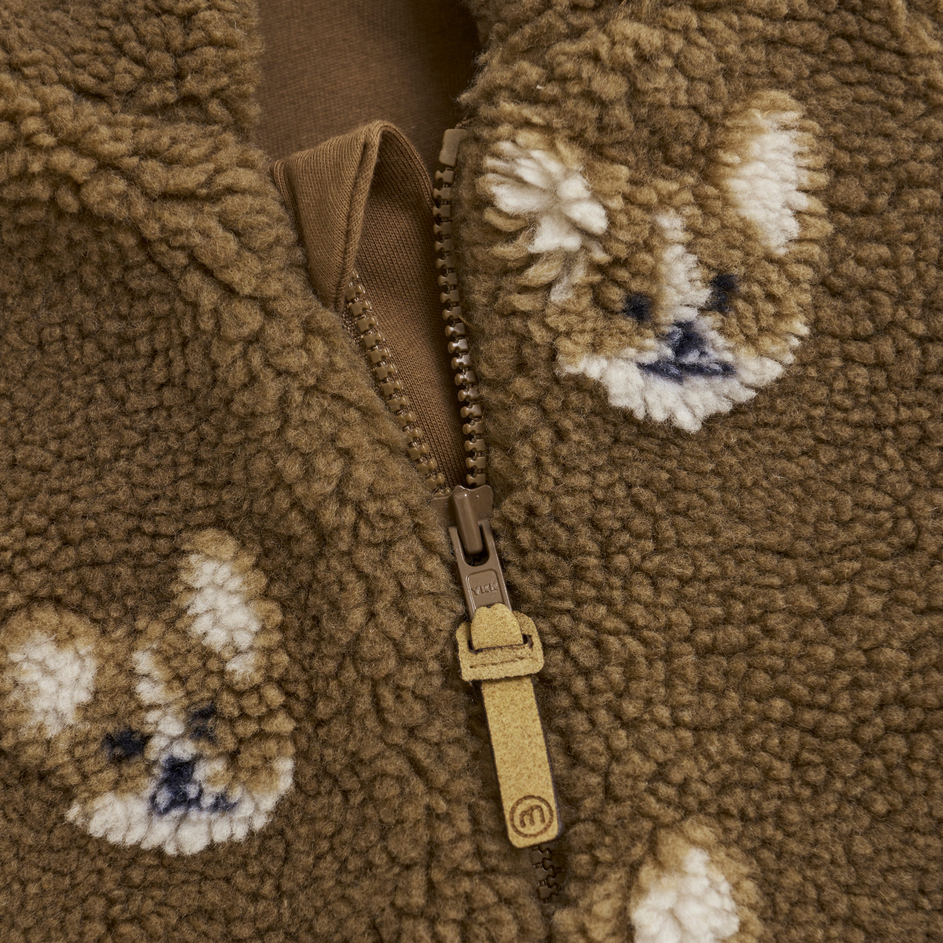Nahaufnahme einer braunen Minymo Jacke mit Reißverschluss in der Mitte. Der Stoff hat eine weiche, flauschige Struktur und ist mit kleinen weißen Hundegesichtern bedruckt, die mit blauen Details über das Material verstreut sind.