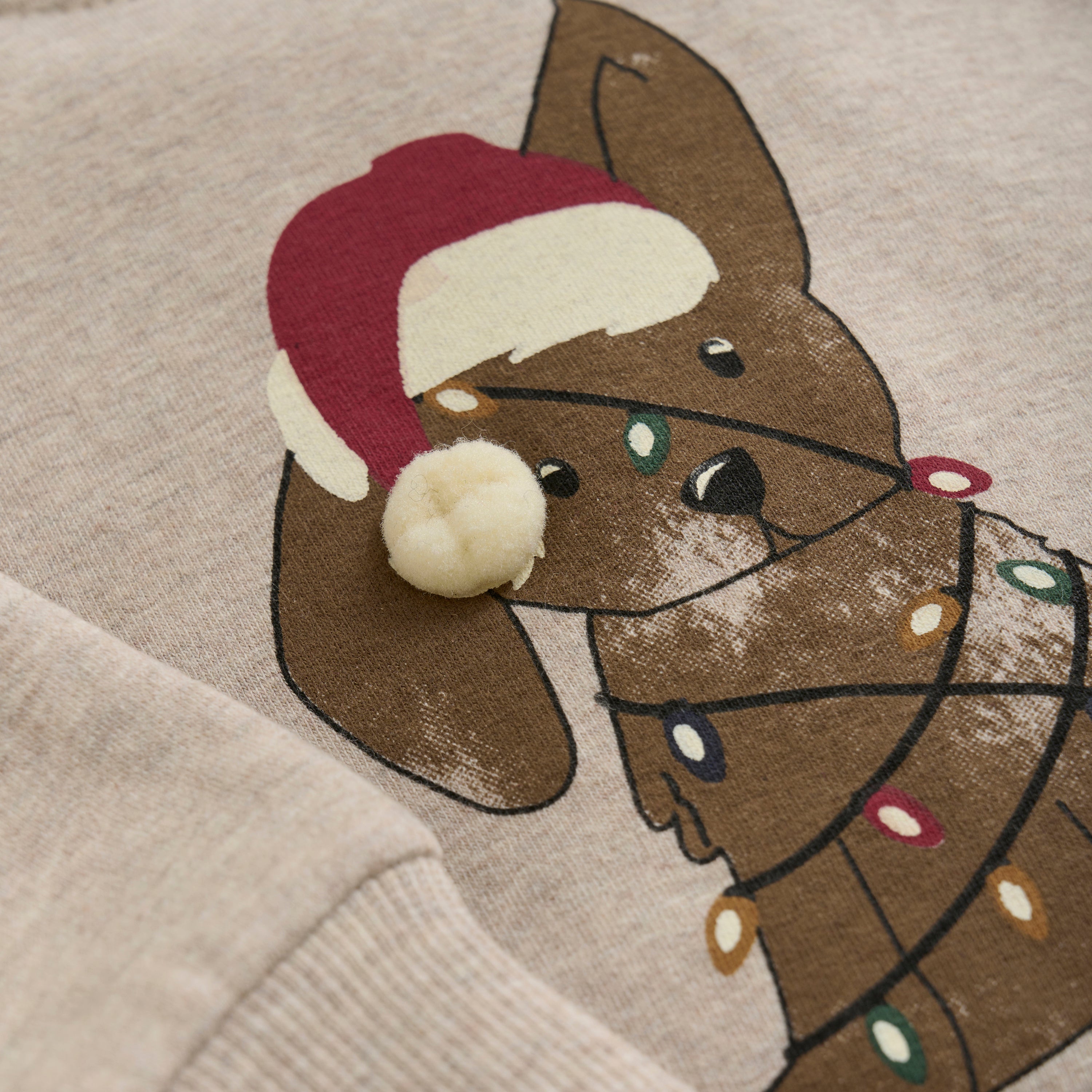 Nahaufnahme eines beigen Minymo-Pullovers mit einer festlichen Illustration eines braunen Hundes mit einer rot-weißen Weihnachtsmannmütze. Der Hund ist mit bunten Weihnachtslichtern geschmückt und wirkt verspielt und fröhlich, was ein entzückendes Weihnachtsdruck-Design mit Feiertagsthema ergibt.