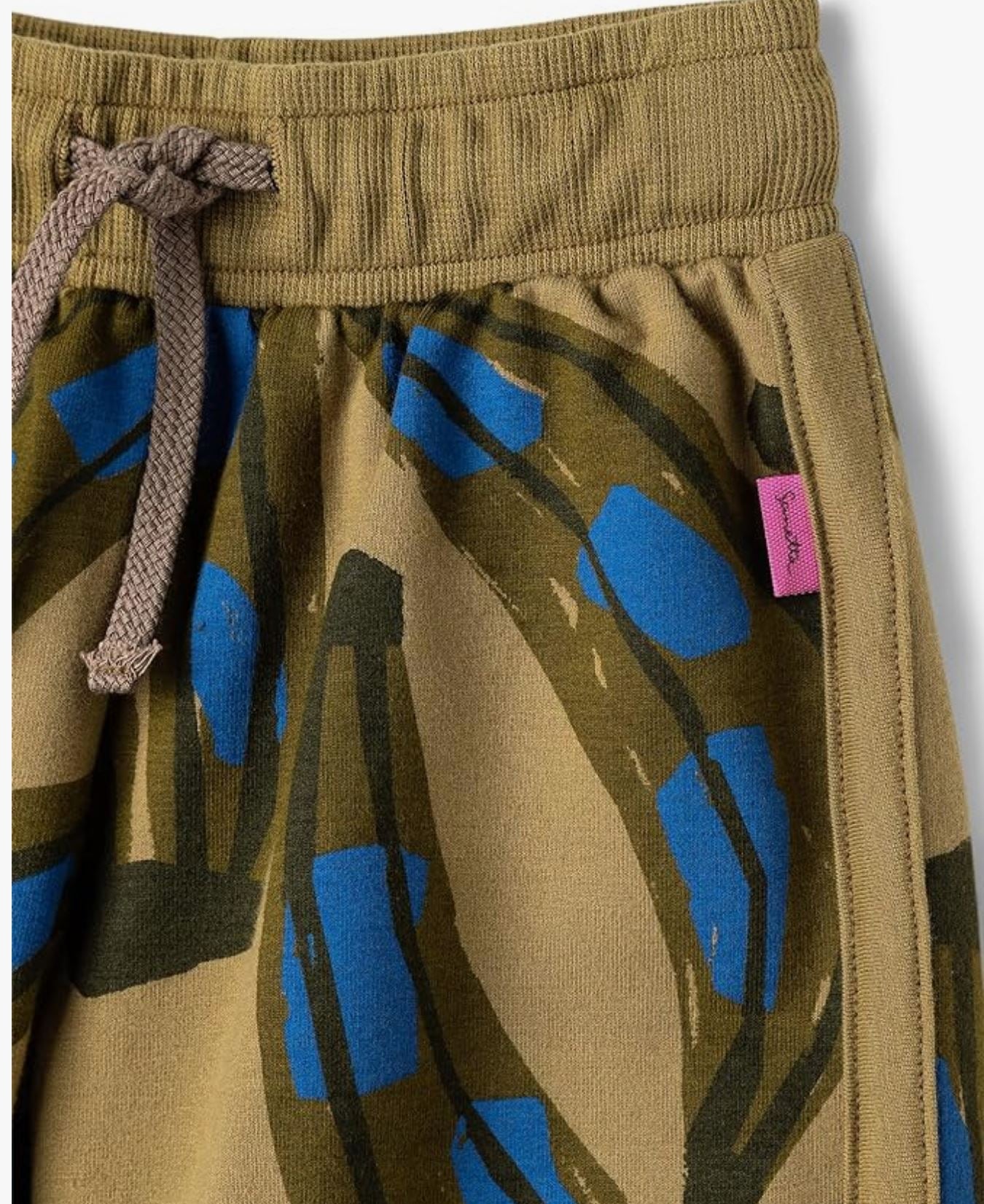 Nahaufnahme der Sanetta - Hose von Sanetta mit Kordelzugbund aus braunem Stoff. Die Hose aus GOTS-zertifizierter Bio-Baumwolle zeigt ein abstraktes Muster in Blau und Grün und ist an der rechten Seitennaht mit einem kleinen rosa Label versehen.