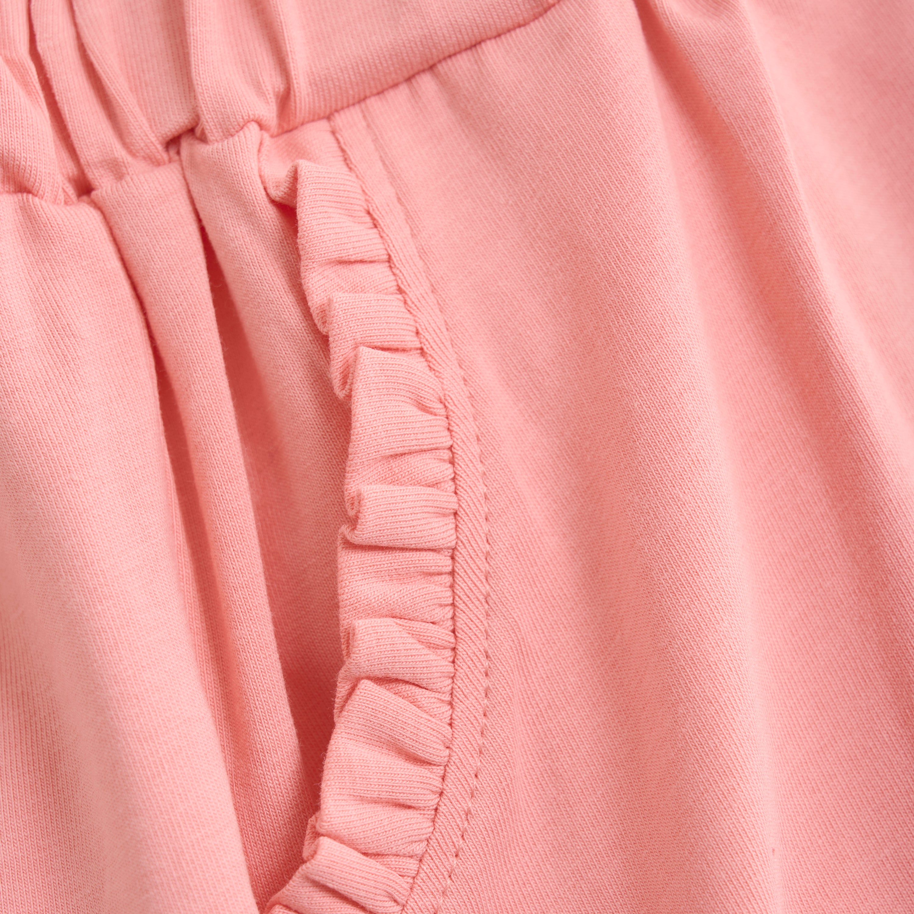 Nahaufnahme der Minymo - Kurze Hosen, die einen rosa Stoff mit gerafftem, elastischem Bund und Rüschenbesatz entlang der Taschenkante zeigt. Die weiche Textur lässt darauf schließen, dass diese Shorts aus bequemem, dehnbarem Baumwollmischgewebe gefertigt sind.