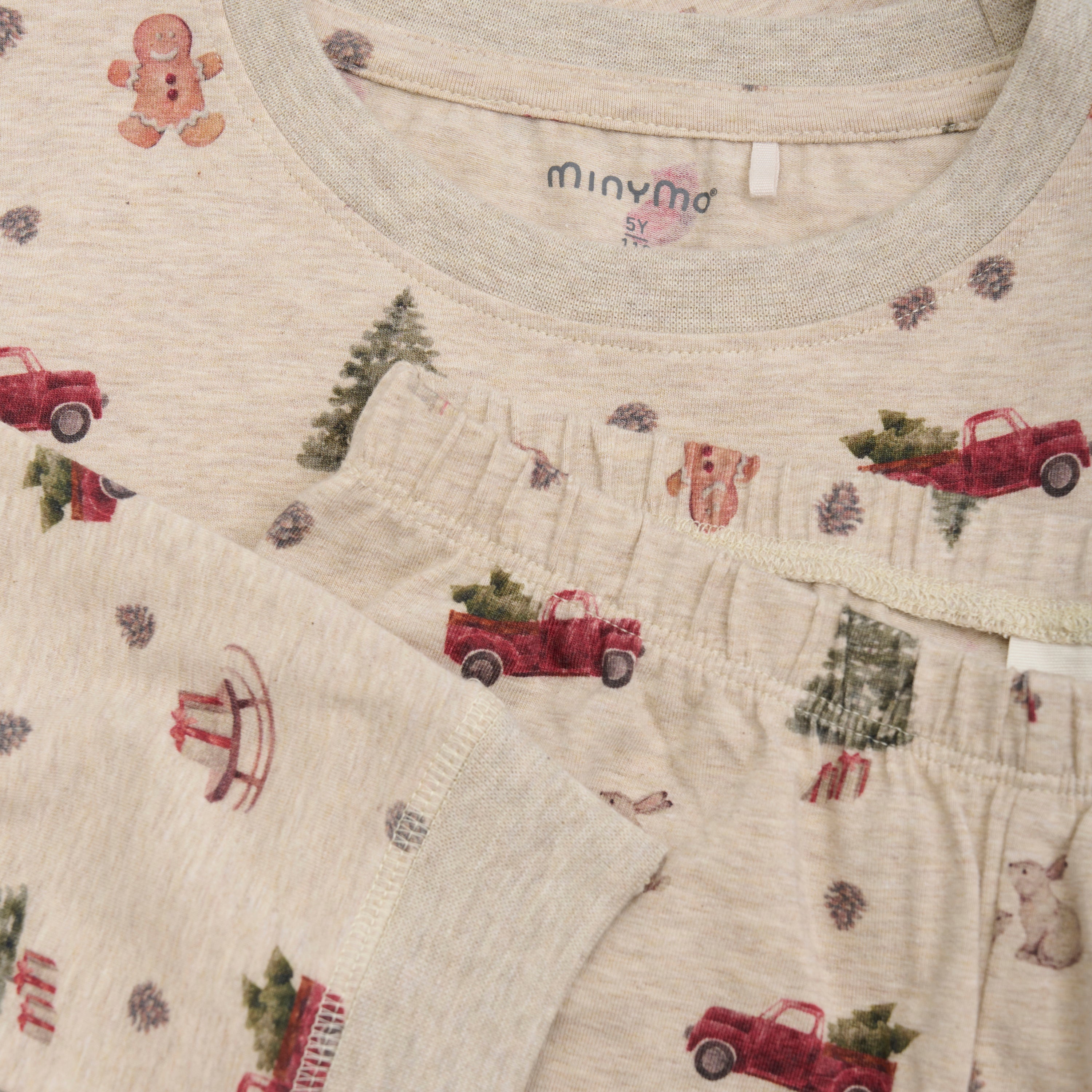 Eine Nahaufnahme des cremefarbenen Minymo-Kinderpyjamas mit festlichem Weihnachtsdruck. Das Nachtwäscheset zeigt rote Lastwagen, die Weihnachtsbäume, Lebkuchenmänner, Bäume und Geschenke transportieren. Auf dem Etikett steht „Minymo, 5 Jahre“. Der Stoff wirkt weich und gemütlich.
