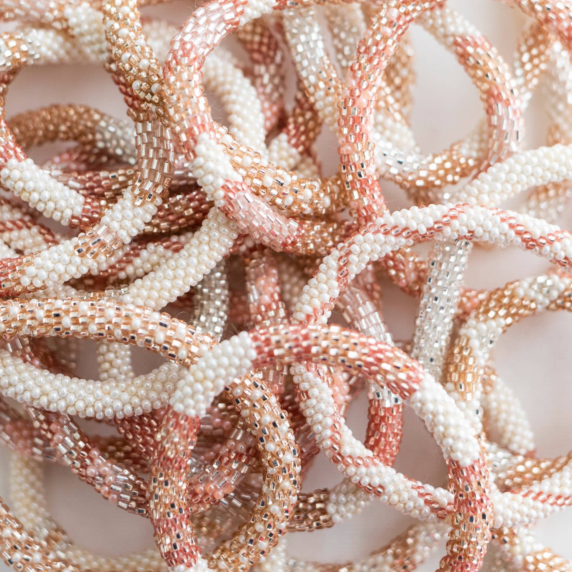 Mehrere kunstvoll mit Perlen verzierte Aid Through Trade - Armbänder Damen in Weiß-, Rosa- und Goldtönen sind ineinander verschlungen. Die kleinen Perlen bilden abwechslungsreiche Muster und verleihen den handgefertigten Armbändern Struktur und optisches Interesse. Sie sind in einer dichten, überlappenden Anordnung aufeinander gestapelt.