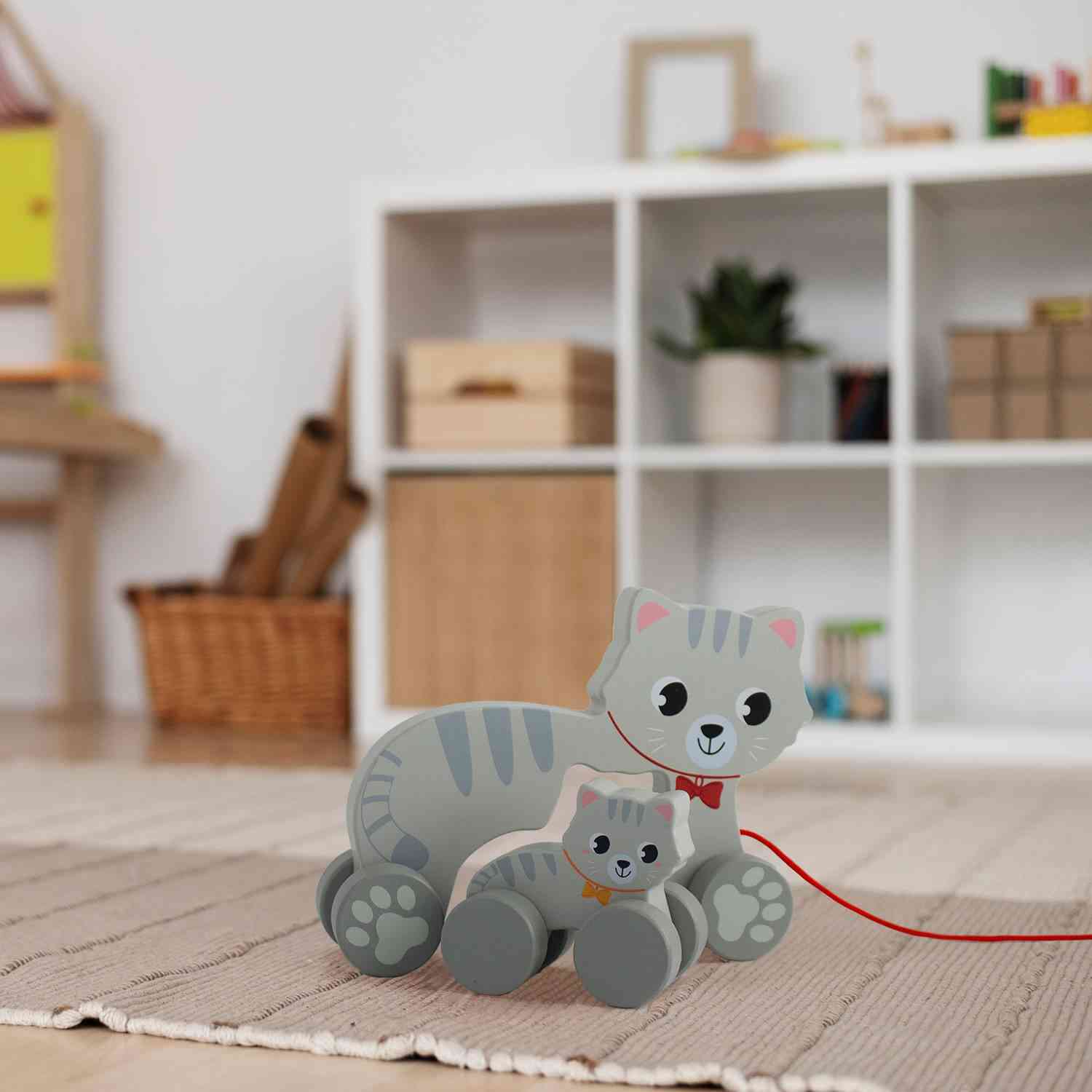 In einem Wohnzimmer stehen zwei süße, passende Ulysse Couleurs d'Enfance - Nachziehtier Katze von Ulysse Couleurs d'Enfance, eine größere und eine kleinere, beide mit grauen Streifen und roten Schleifen. Perfekte Spielsachen für Zweijährige, sie stehen auf einem gewebten Teppich, mit einem weißen Regal, einer Topfpflanze und einigen Aufbewahrungsboxen im Hintergrund.