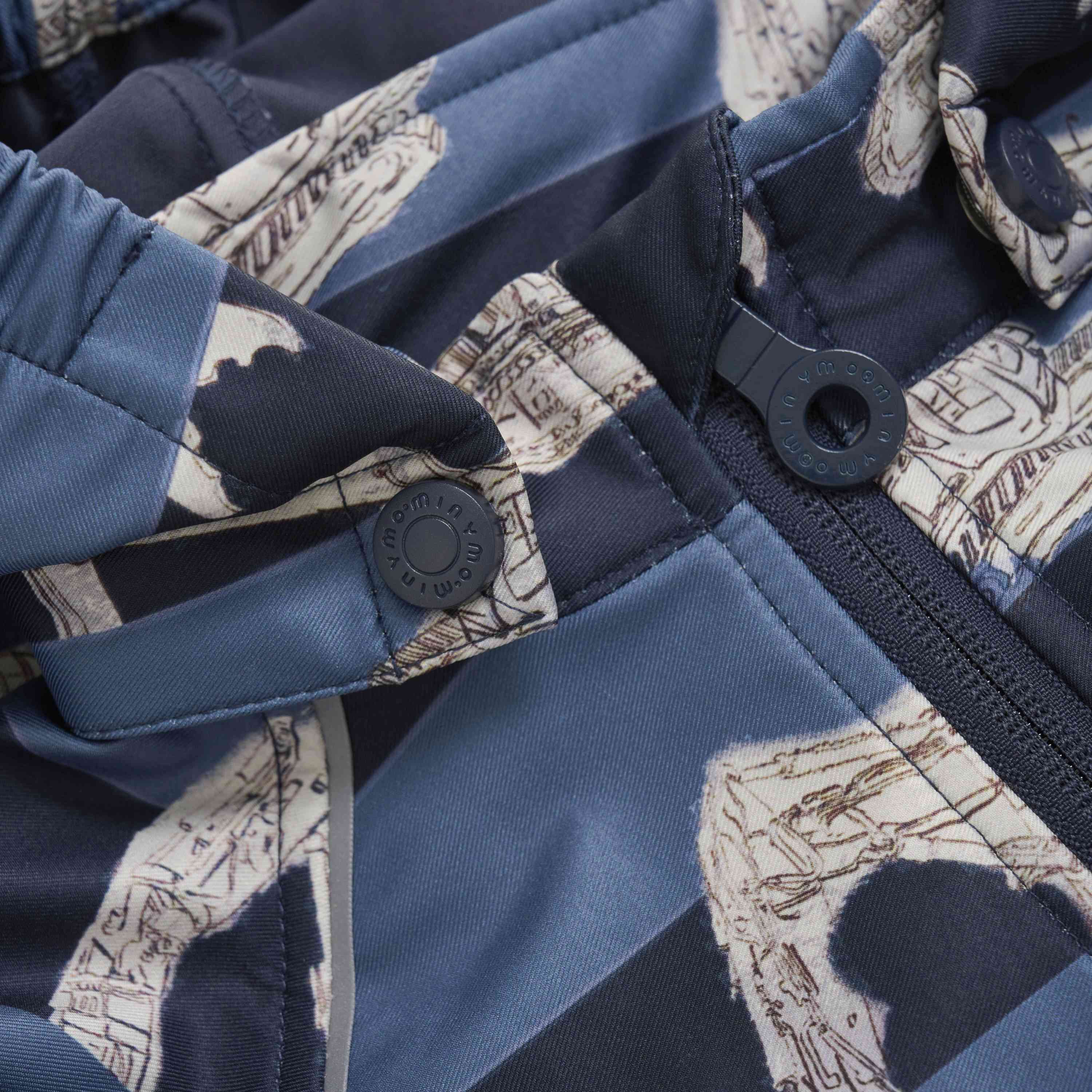 Nahaufnahme eines GOTS-zertifizierten blau-beigen Minymo - Softshell-Anzugs von Minymo mit Reißverschluss vorne und Knopfdetails. Der Stoff hat ein gemustertes Design mit dunkel- und hellblauen Abschnitten und beigen Illustrationen. Die Knöpfe sind ideal für Waldspielgruppenkleider und haben einen eingeprägten Markentext.