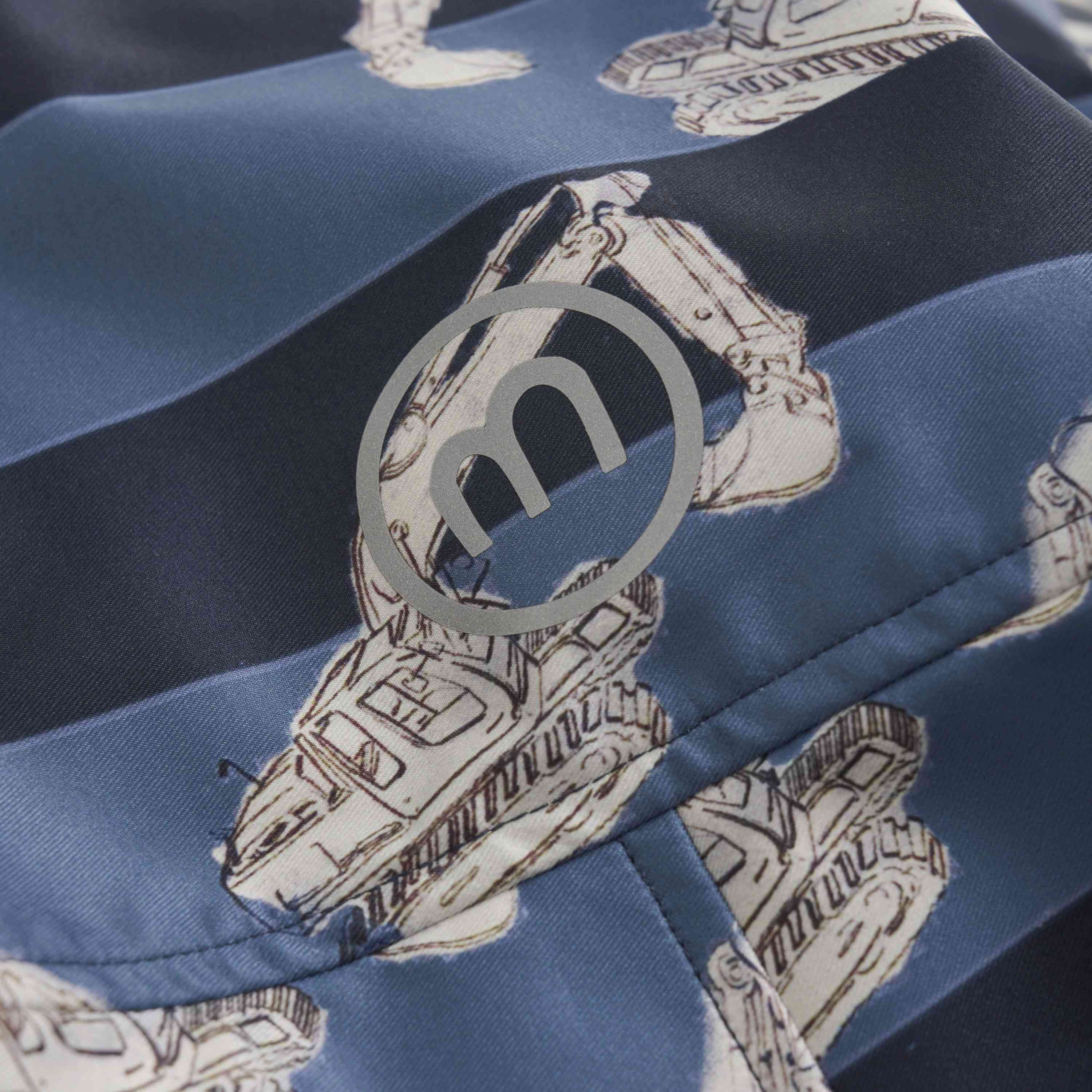 Nahaufnahme eines Stoffes mit einem Muster aus weißen Baggern auf einem blau gestreiften Hintergrund. Auf der Oberfläche ist ein aufgedrucktes kreisförmiges Logo mit einem stilisierten „m“ sichtbar, das darauf hinweist, dass der Stoff GOTS-zertifiziert ist. Die Textur des Stoffes Minymo - Softshell-Anzug wirkt glatt und seidig.