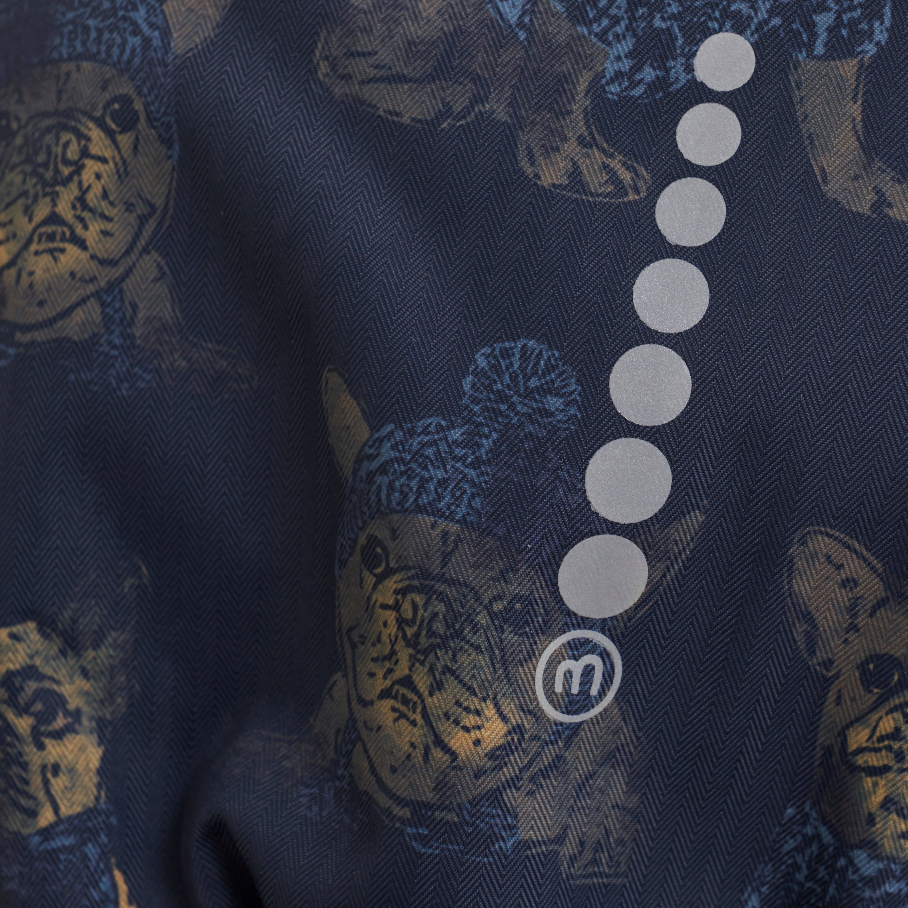 Nahaufnahme eines gemusterten Stoffes mit Katzenabbildungen mit blauen Strickmützen. Der dunkelblaue Stoff eignet sich perfekt für die Herstellung eines wärmeren Minymo-Schneeanzugs von Minymo und besticht durch ein dezentes Fischgrätenmuster. Hellgraue kreisförmige Motive sind auf der rechten Seite vertikal ausgerichtet, mit einem kleinen kreisförmigen Monogramm am unteren Rand.