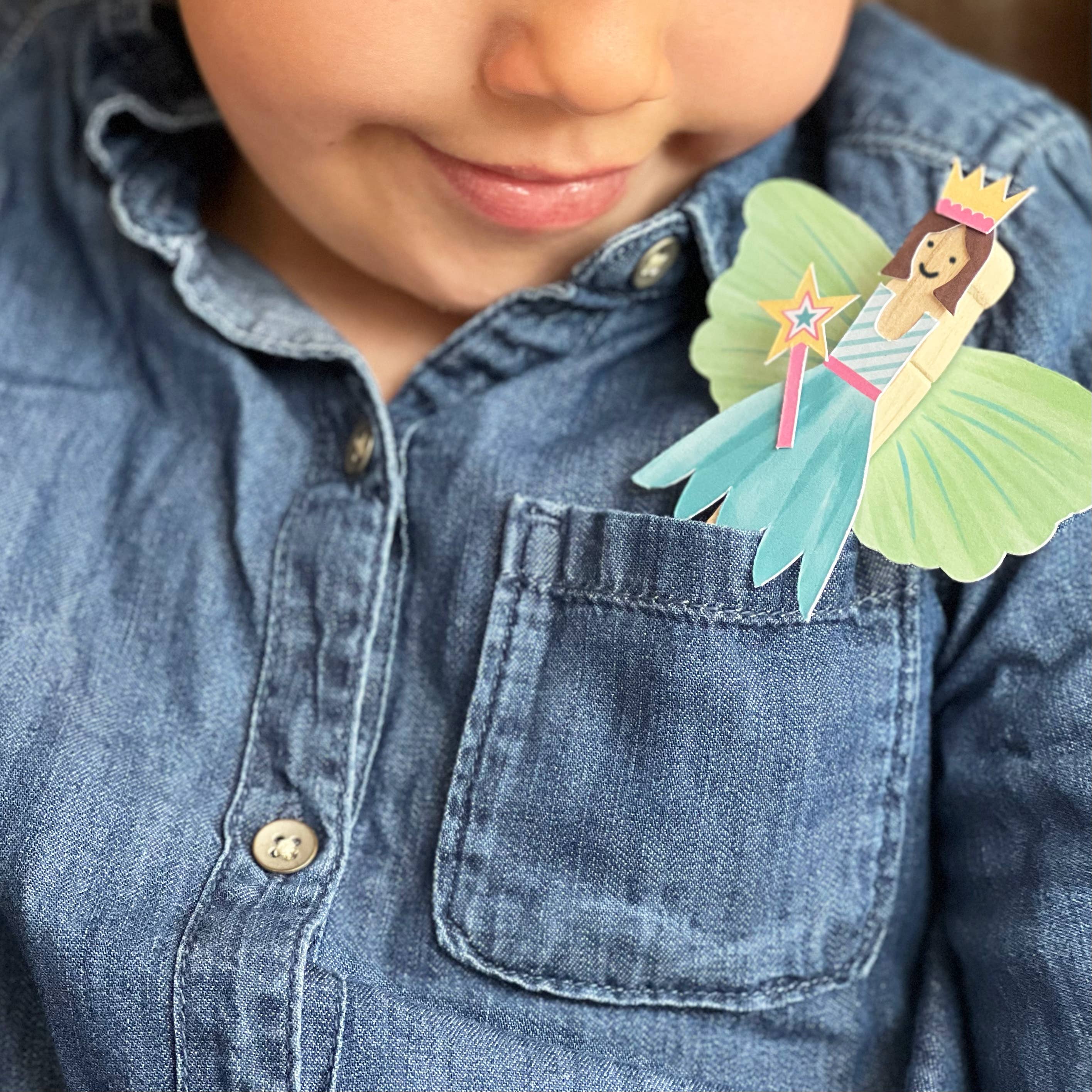 Ein Kind in einem blauen Jeanshemd lächelt sanft. Ein bunter Papierausschnitt einer Feenprinzessin mit Krone und Flügeln aus der Cotton Twist - DIY Fairy Pog-Puppe von Cotton Twist steckt in der Vordertasche des Hemdes. Die aus umweltfreundlichen Materialien gefertigte Fee hält einen Sternenstab und trägt ein blaues Kleid, um die Feinmotorik der Kinder zu fördern.