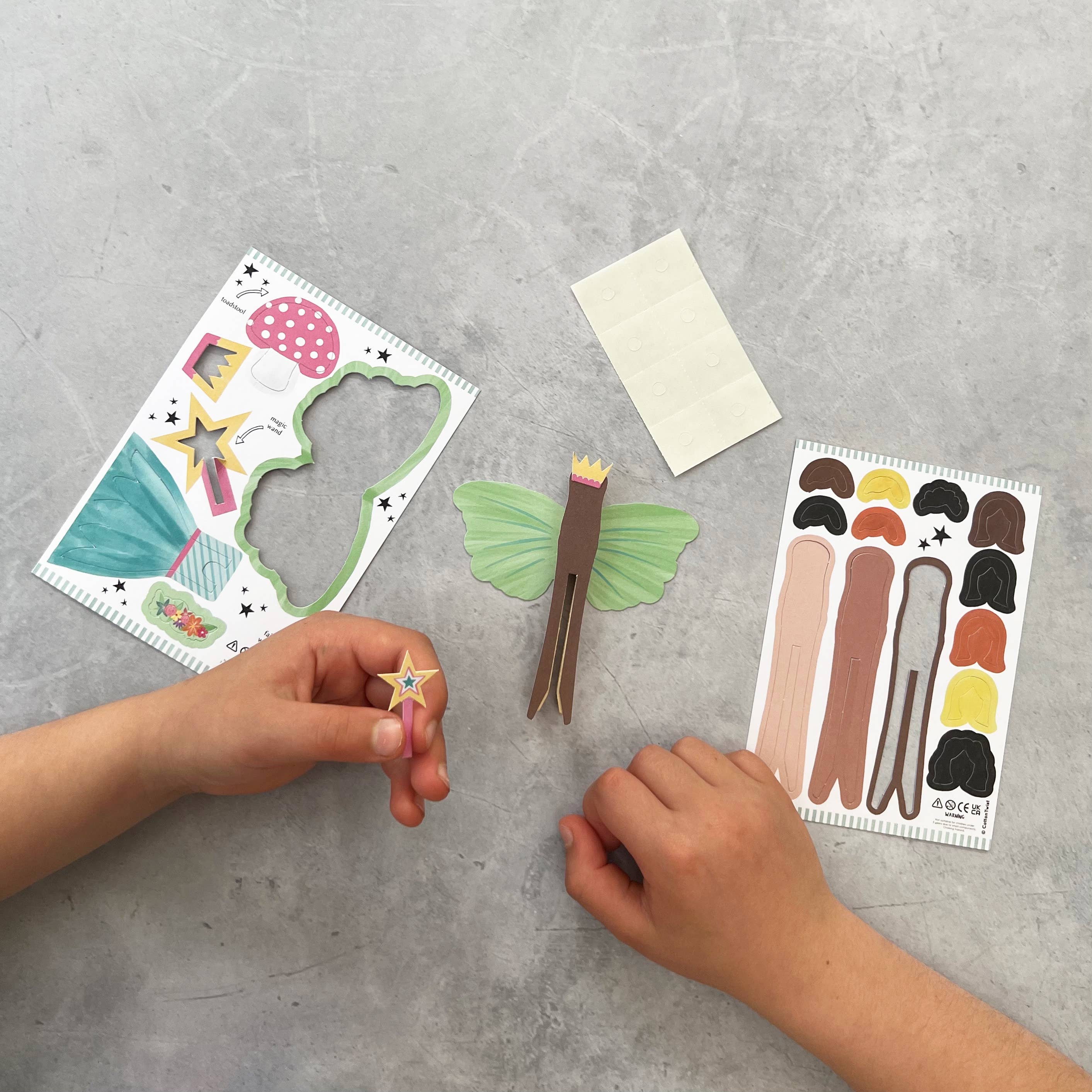 Zwei Hände bauen eine Cotton Twist - DIY Fairy Pog-Puppe auf einer hellgrauen Oberfläche zusammen, wobei umweltfreundliche Materialien verwendet werden. Das Projekt umfasst Aufkleber, Papierausschnitte in verschiedenen Formen und Farben sowie Klebestücke. Eine Hand hält einen kleinen sternförmigen Gegenstand, während die andere einen grünen Feenausschnitt mit einer Krone hält. Diese Aktivität fördert die Feinmotorik.