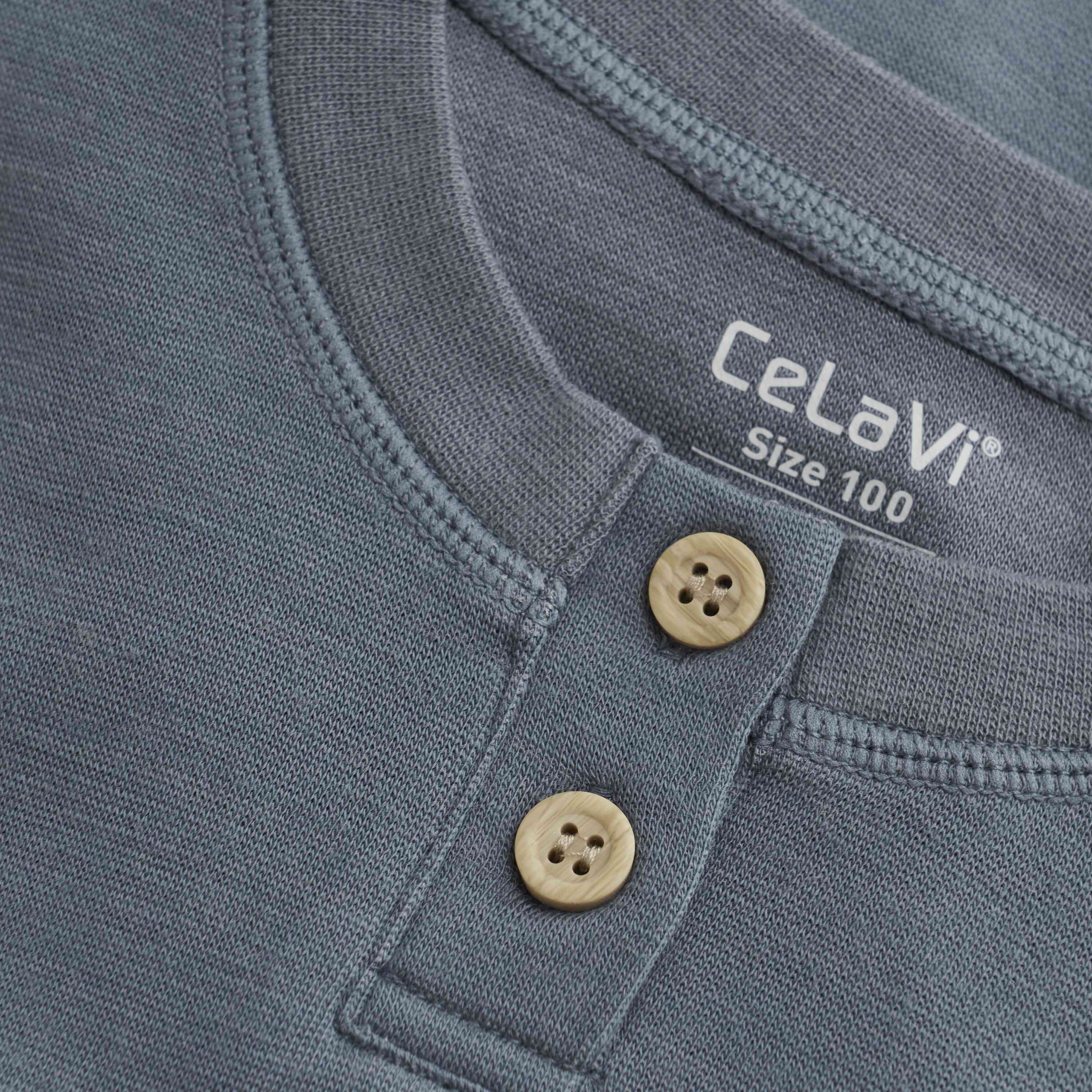 Nahaufnahme einer blaugrauen CeLaVi Bluse mit Rundhalsausschnitt, zwei beigen Knöpfen unter dem Kragen und einem sichtbaren Etikett mit der Marke CeLaVi und der Größe 100. Der Stoff hat eine weiche, strukturierte Optik und erfüllt die Qualitätskriterien des OEKO-TEX STANDARD 100.