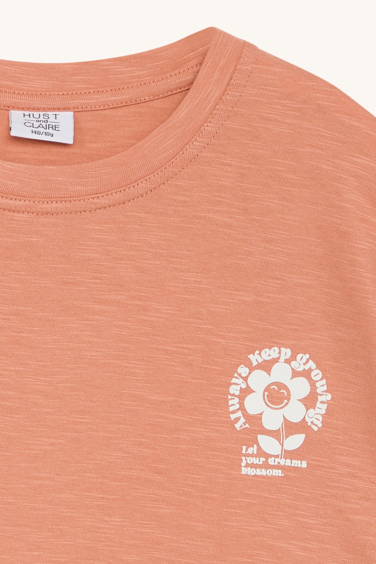 Foto eines orangefarbenen Hust and Claire - Agne T-Shirts mit einer weißen Blumengrafik auf der Vorderseite. Die Blume hat ein lächelndes Gesicht und der Text darum lautet: „Wachse immer weiter“ und „Lass deine Träume erblühen.“ Dieses Kinderartikelgeschäft in Brimensdorf verkauft mitwachsende Shirts wie dieses von Hust & Claire, Größe 104/4y.