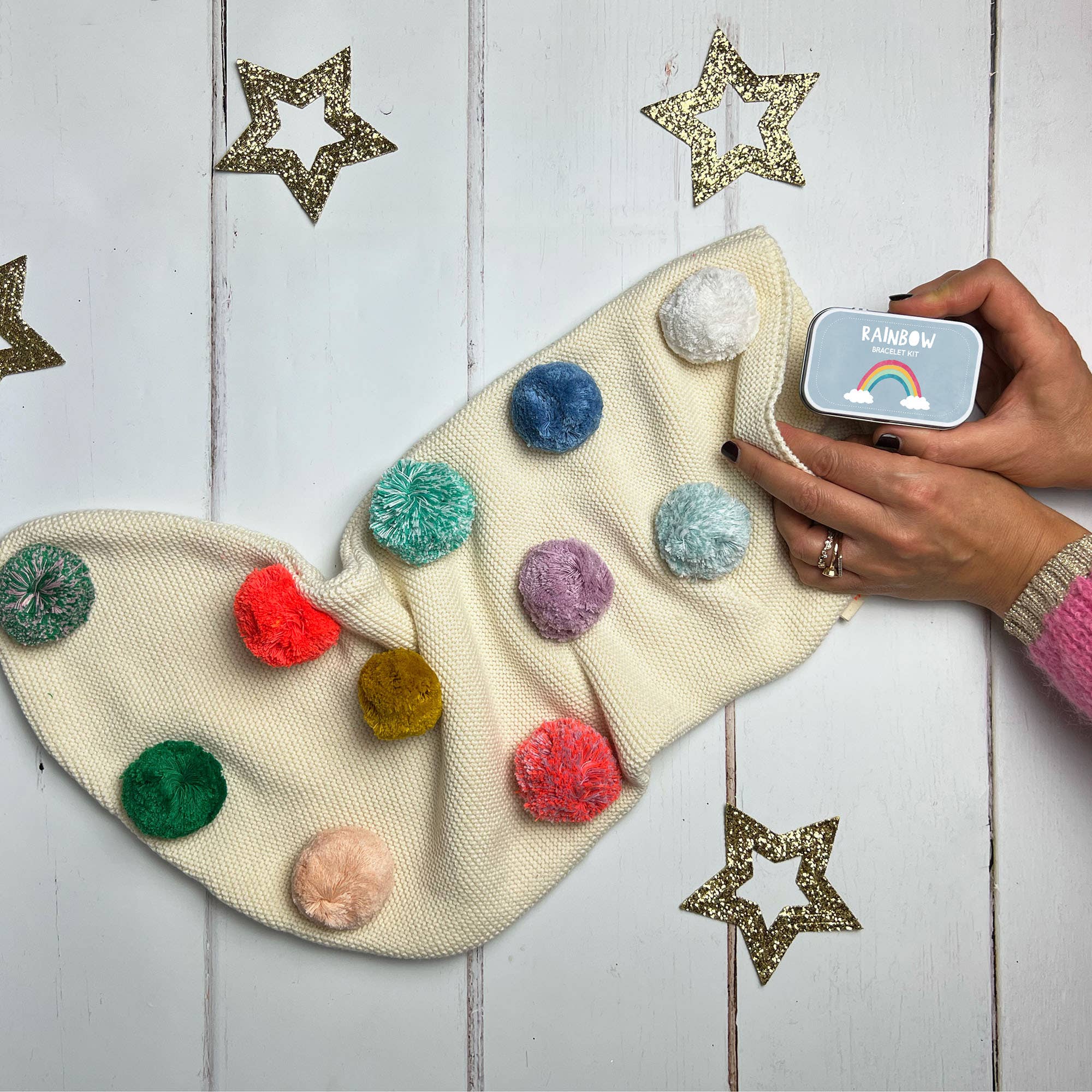 Eine Hand, die ein Cotton Twist – DIY Regenbogen-Armband neben einer cremefarbenen Socke mit bunten Pompons hält, vor einem hölzernen Hintergrund mit goldenen sternförmigen Verzierungen, lässt auf lebendige Gastgeschenke für jede Feier schließen.