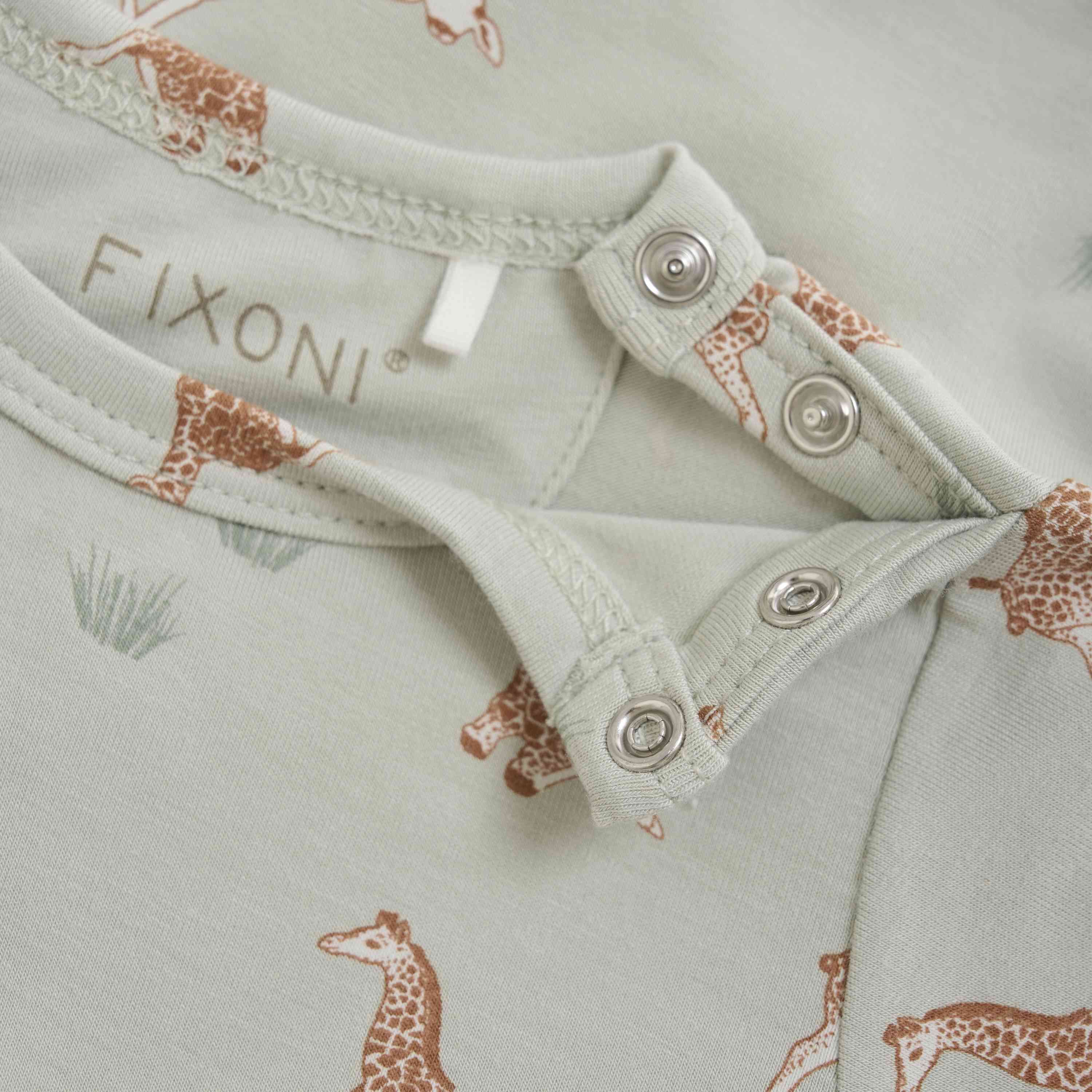 Nahaufnahme eines hellgrünen Baby-Stramplers mit einem Muster aus kleinen braunen Giraffen und Pflanzen. Der Fixoni - Body kurzarm verfügt über einen Druckknopfverschluss in der Nähe des Halsausschnitts und das Stoffetikett zeigt „Fixoni“.