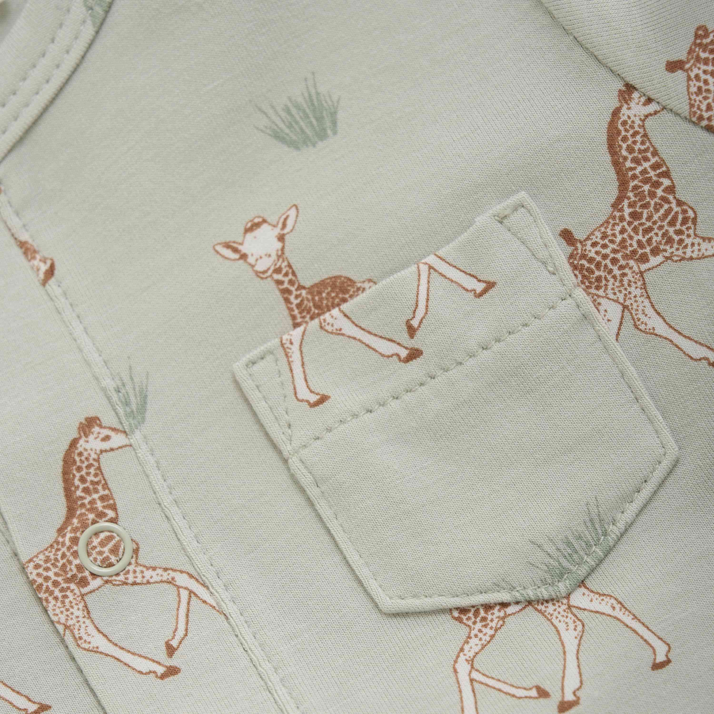 Nahaufnahme eines hellgrünen Stoffes mit einer Tasche. Der GOTS-zertifizierte Stoff ist mit kleinen Bildern von braunen und weißen Giraffenbabys und Grasbüscheln gemustert. Die Tasche ist auf den Fixoni - Stampler Langarm von Fixoni aufgenäht und fügt sich nahtlos in das Hintergrunddesign ein.