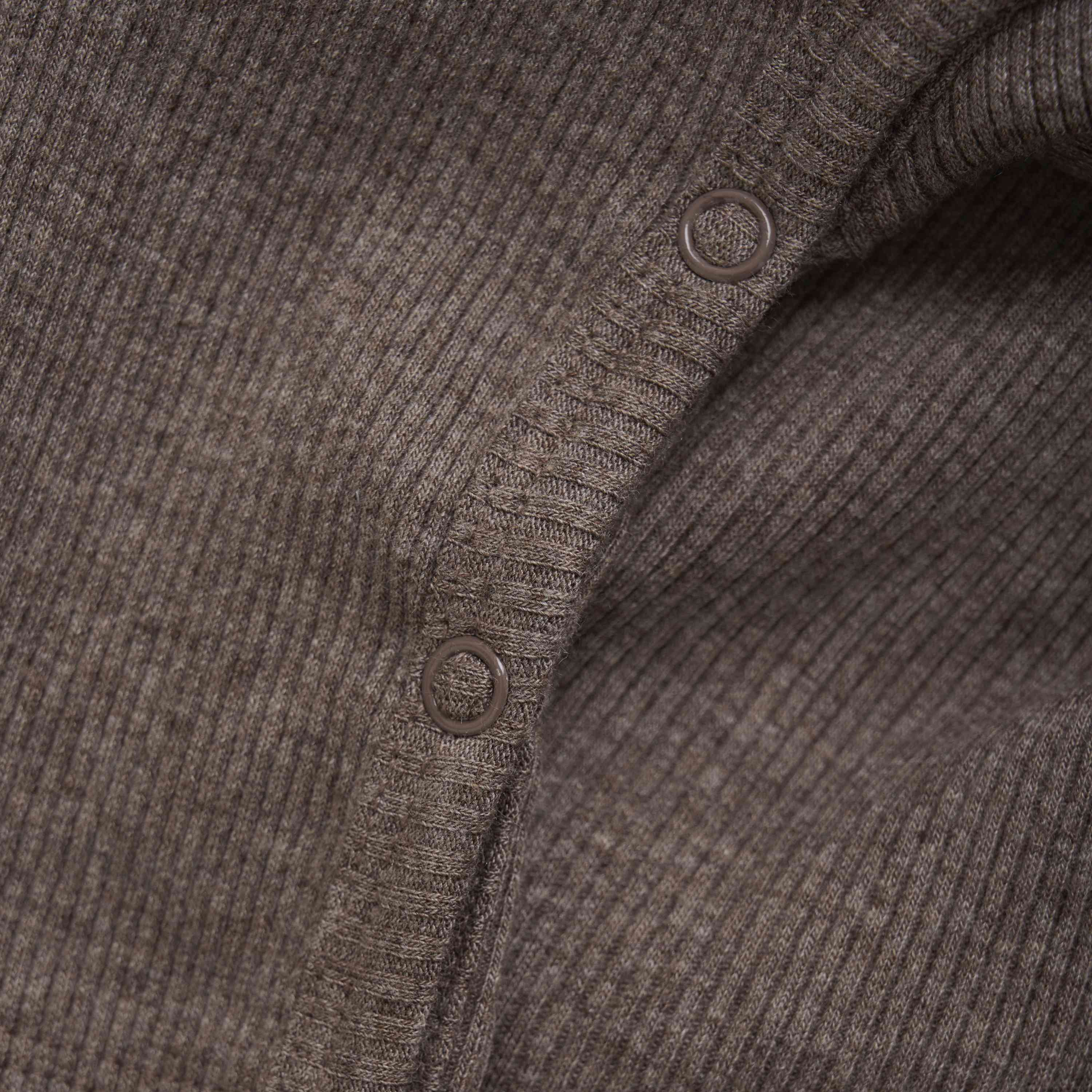 Nahaufnahme eines strukturierten braunen Stoffes mit gerippten Details und verstärkten Knopflöchern, wie er beim Fixoni - Strampler von Fixoni zu sehen ist. Der Fokus liegt auf dem komplizierten Strickmuster und den dunkelbraunen TENCEL™-Fäden, die die Qualität und Webart des Materials hervorheben.