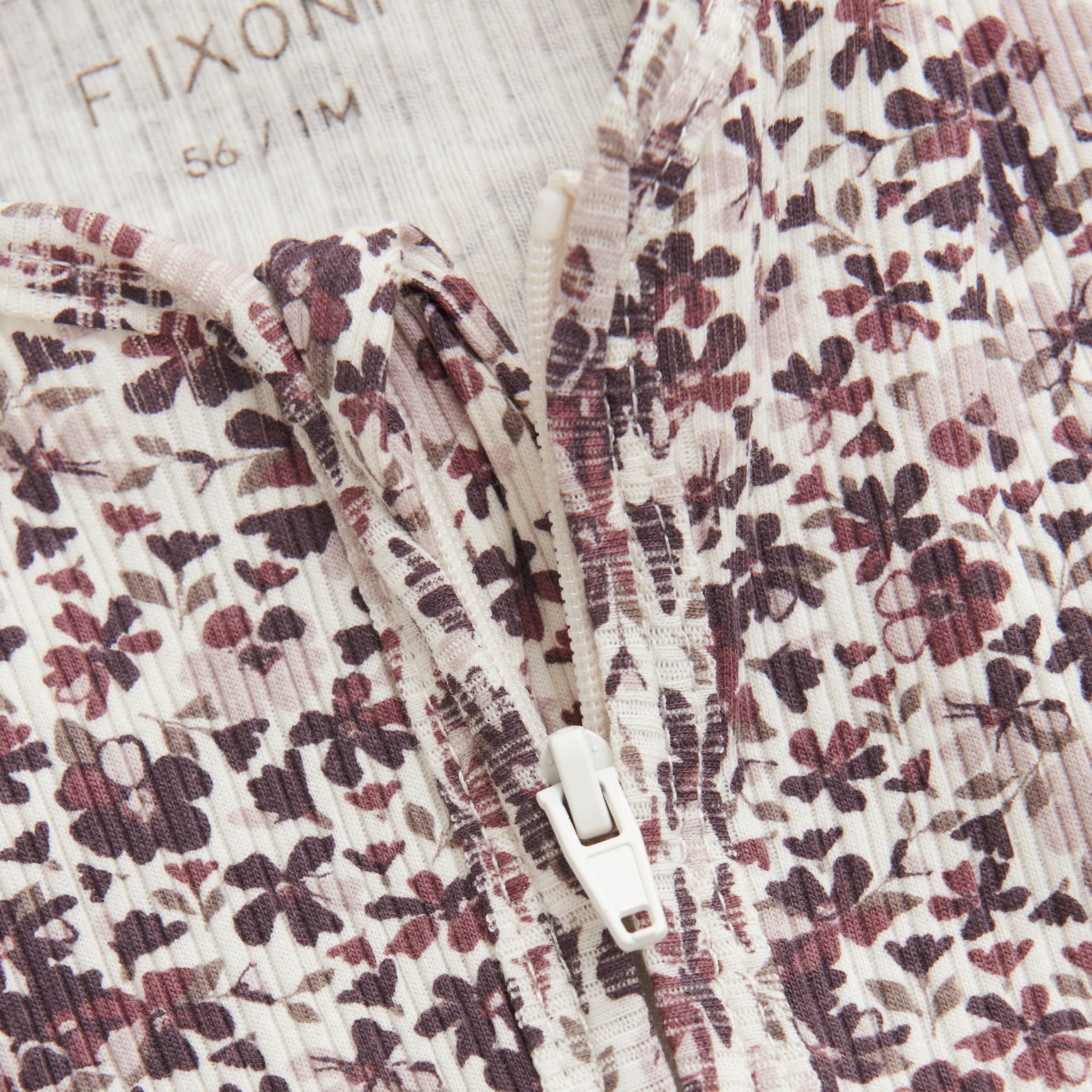 Nahaufnahme eines Stoffes mit Blumenmuster in gedeckten Farben, mit teilweise geöffnetem Reißverschluss. Das Stoffetikett der Marke „Fixoni“ und der Größe „56 1M“ ist in der oberen linken Ecke sichtbar. Dieses zarte Design, ideal für einen Fixoni-Pyjama, umfasst kleine Blumen in Braun- und Rosatönen.