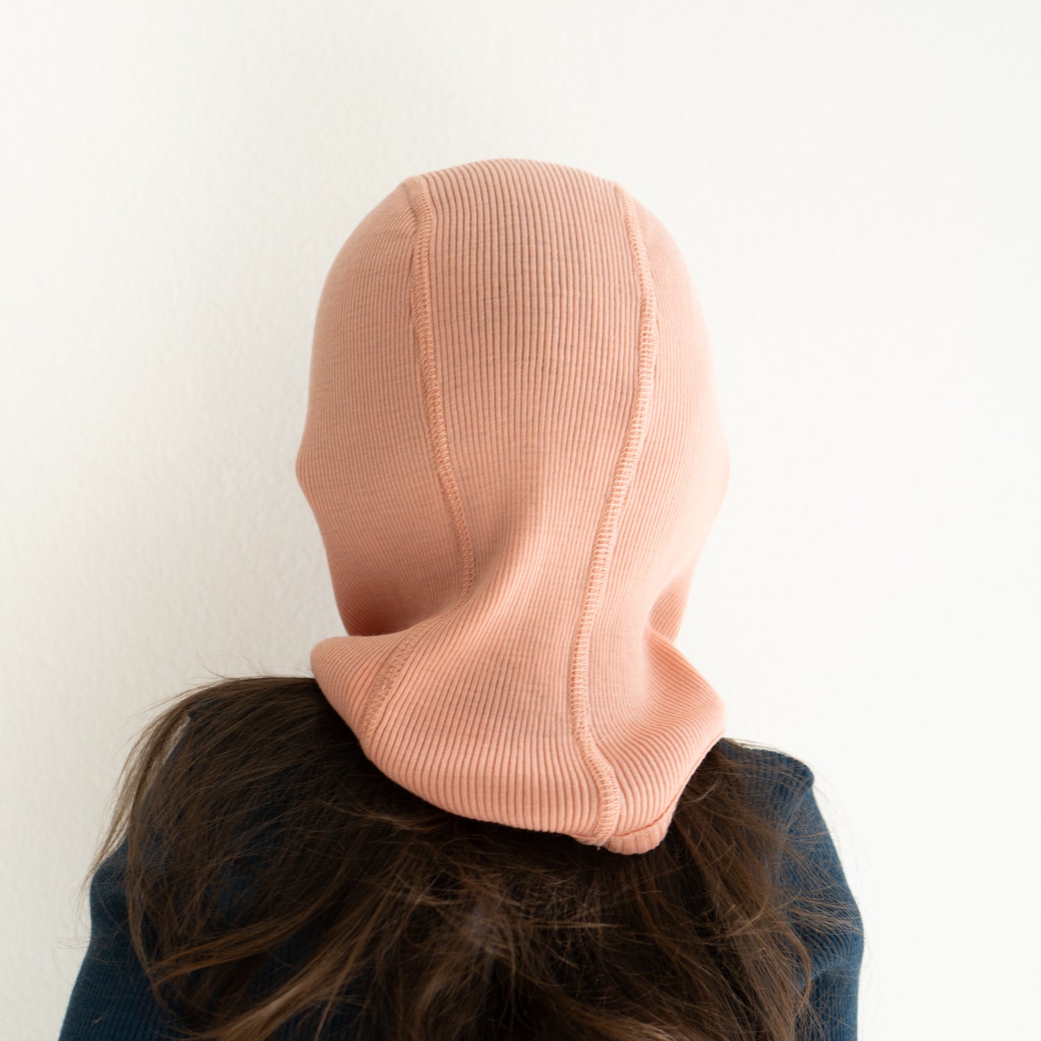 Person mit langen braunen Haaren, die die Minimalisma-Mütze trägt, eine hellrosa gerippte Sturmhaube aus Merinowolle von Minimalisma, von hinten betrachtet vor einem schlichten weißen Hintergrund.