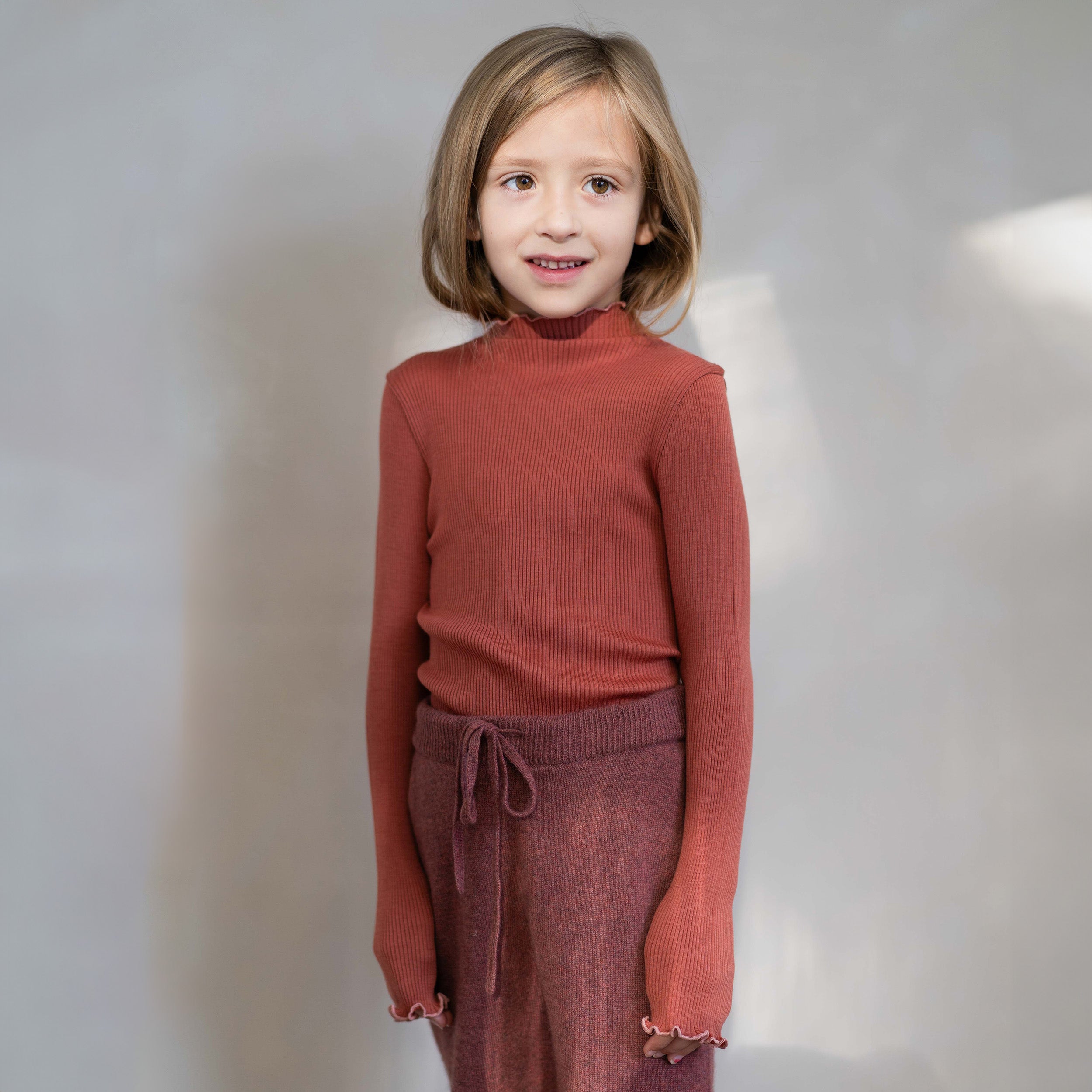 Ein kleines Kind mit schulterlangem blondem Haar steht vor einem schlichten Hintergrund, gekleidet in das Langarmshirt von Minimalisma und dazu passende hoch taillierte Hosen mit Kordelzug. Der Responsible Wool Standard stellt sicher, dass ihr rostfarbenes geripptes Merinowoll-Outfit sowohl gemütlich als auch nachhaltig ist. Das Kind lächelt leicht und schaut zur Seite.