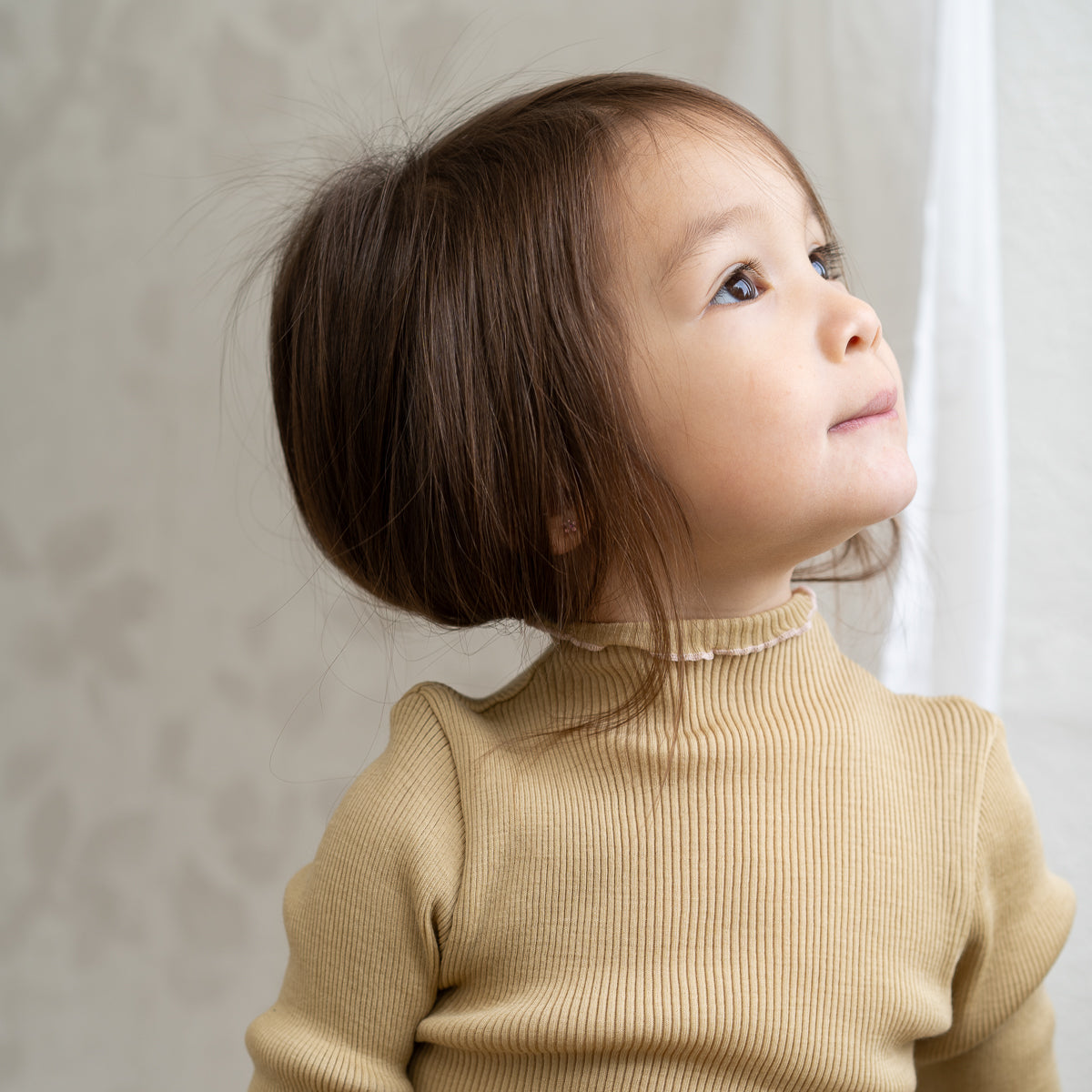 Ein kleines Kind mit schulterlangem braunem Haar blickt nachdenklich nach oben. Es trägt das minimalisma - Langarm Shirt der Marke minimalisma, eine beige, langärmlige gerippte Rollkragenbluse aus Merinowolle. Der Hintergrund ist sanft beleuchtet und zeigt helle, abstrakte Muster an der Wand.