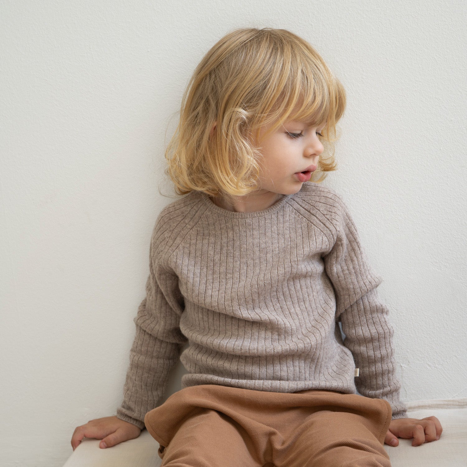 Ein kleines Kind mit blonden, schulterlangen Haaren sitzt vor einer weißen Wand und blickt zur Seite hinunter. Es trägt ein hellbeiges, langärmeliges „minimalisma - Langarm Shirt (Kopie)“ aus Responsible Wool Standard-Materialien und eine braune Hose. Sein Gesichtsausdruck wirkt nachdenklich.