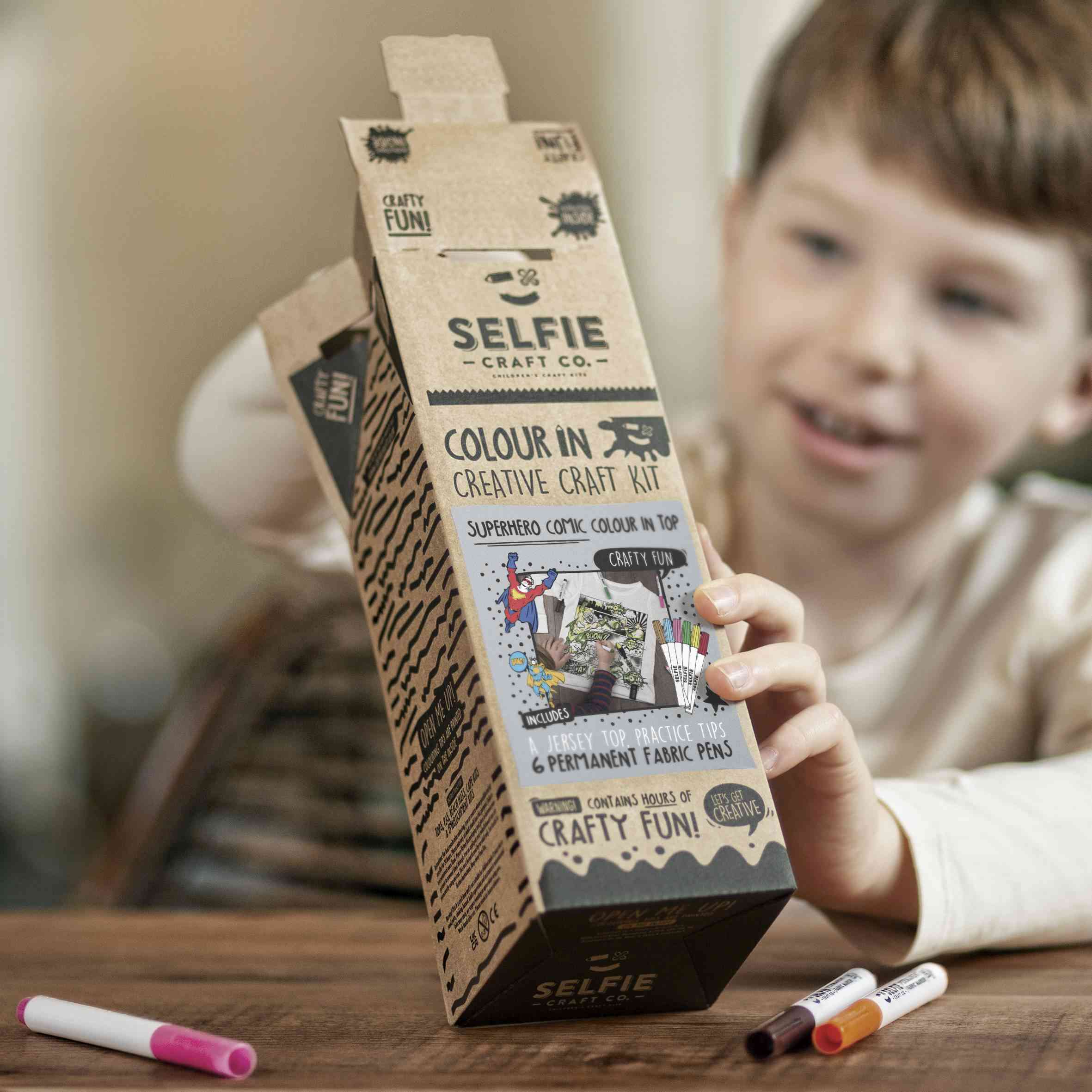 Ein Kind sitzt an einem Tisch und hält ein offenes „Selfie Craft“ Selfie Craft Co. – Superhelden Comic: Oberteil zum Ausmalen. Das Paket enthält leuchtende Illustrationen und einen Text, der den Inhalt beschreibt, darunter auch Stoffmarker. Zwei Marker, einer rosa und einer orange, liegen auf dem Tisch im Vordergrund – perfekt für leichtes basteln mit Kindern.