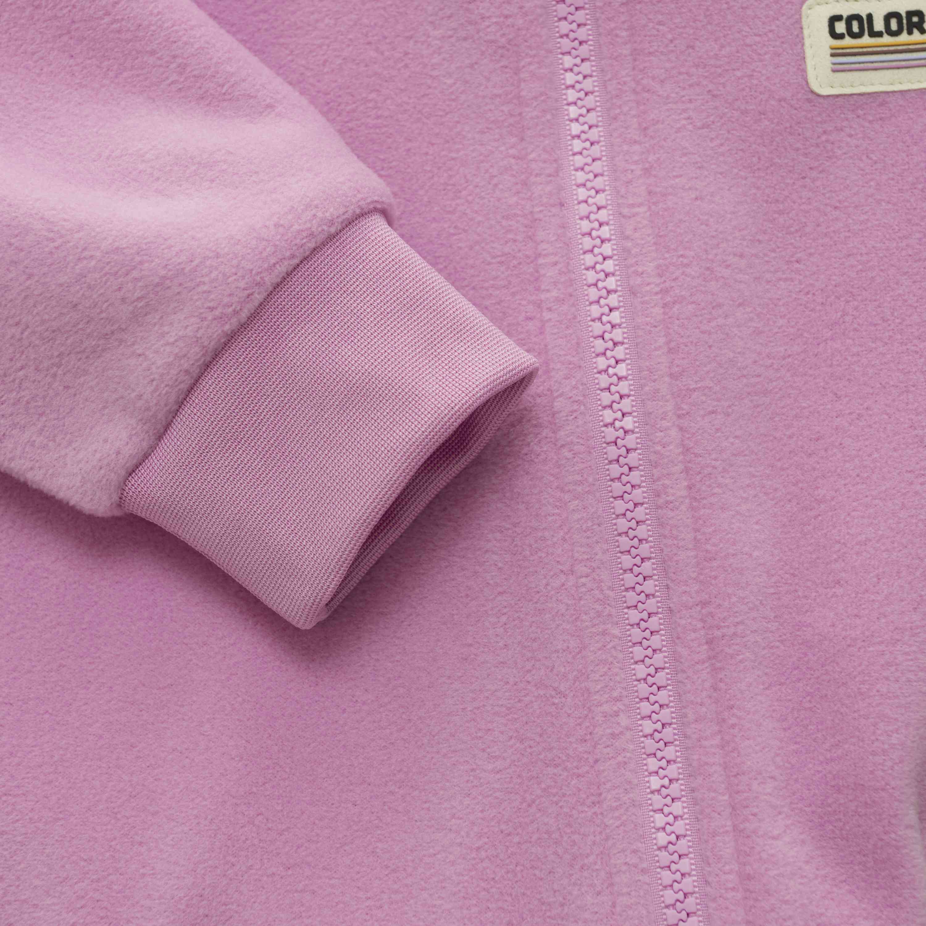 Nahaufnahme eines hellvioletten Baby-Fleeceanzugs von Color Kids mit Reissverschluss und gerippten Bündchen an den Ärmeln. Der Stoff sieht weich aus, wahrscheinlich Fleece. In der Nähe des Reissverschlusses ist ein kleines Etikett mit teilweise sichtbarem „Color Kids“-Schriftzug aufgenäht.