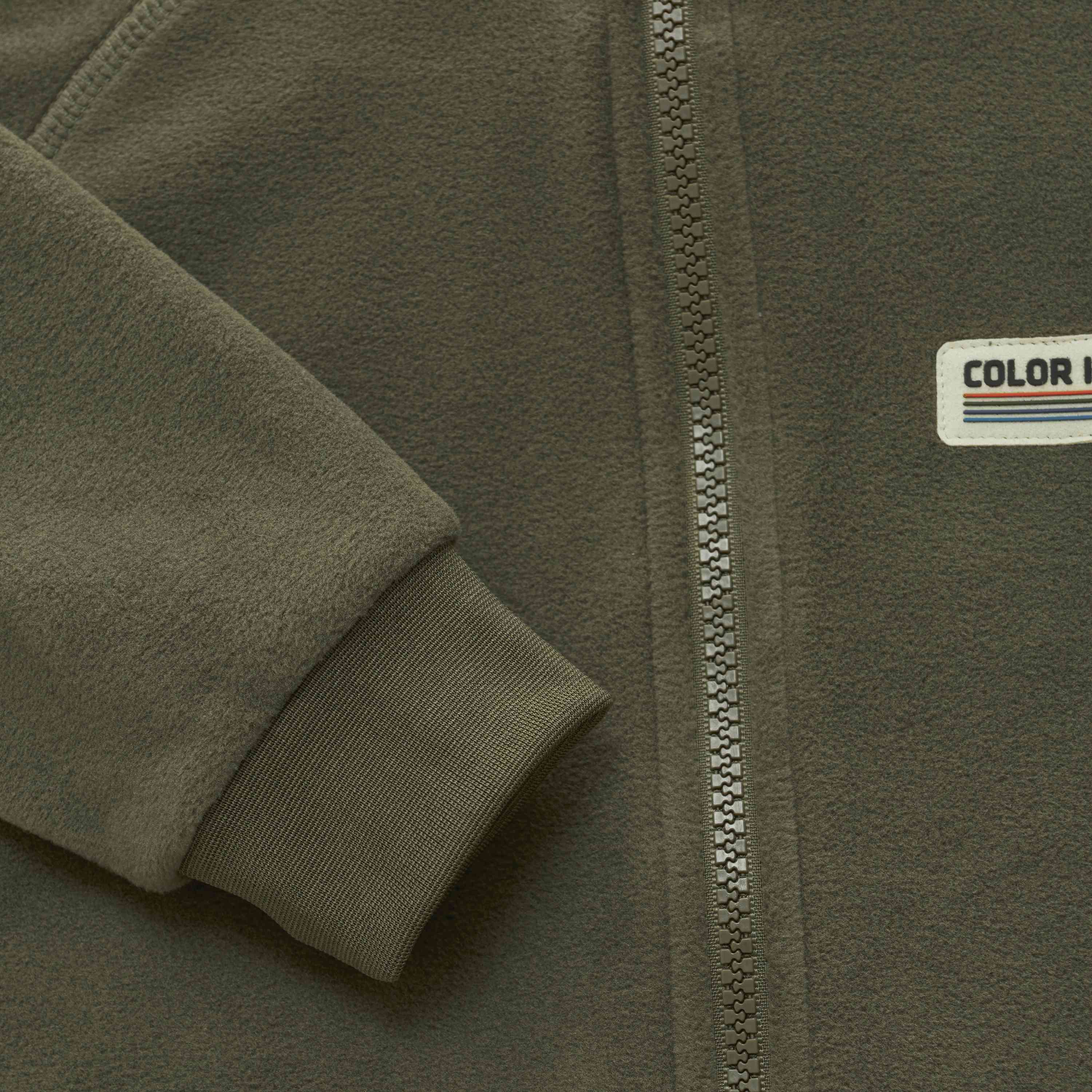Nahaufnahme einer grünen Fleecejacke, auf der der Ärmel, ein Teil des Reissverschlusses und ein Patch mit dem Text „Color Kids“ und einem kleinen Streifenmuster zu sehen sind. Der Baby-Fleeceanzug scheint aus weichem, bequemem Material zu bestehen, das schnell trocknet.