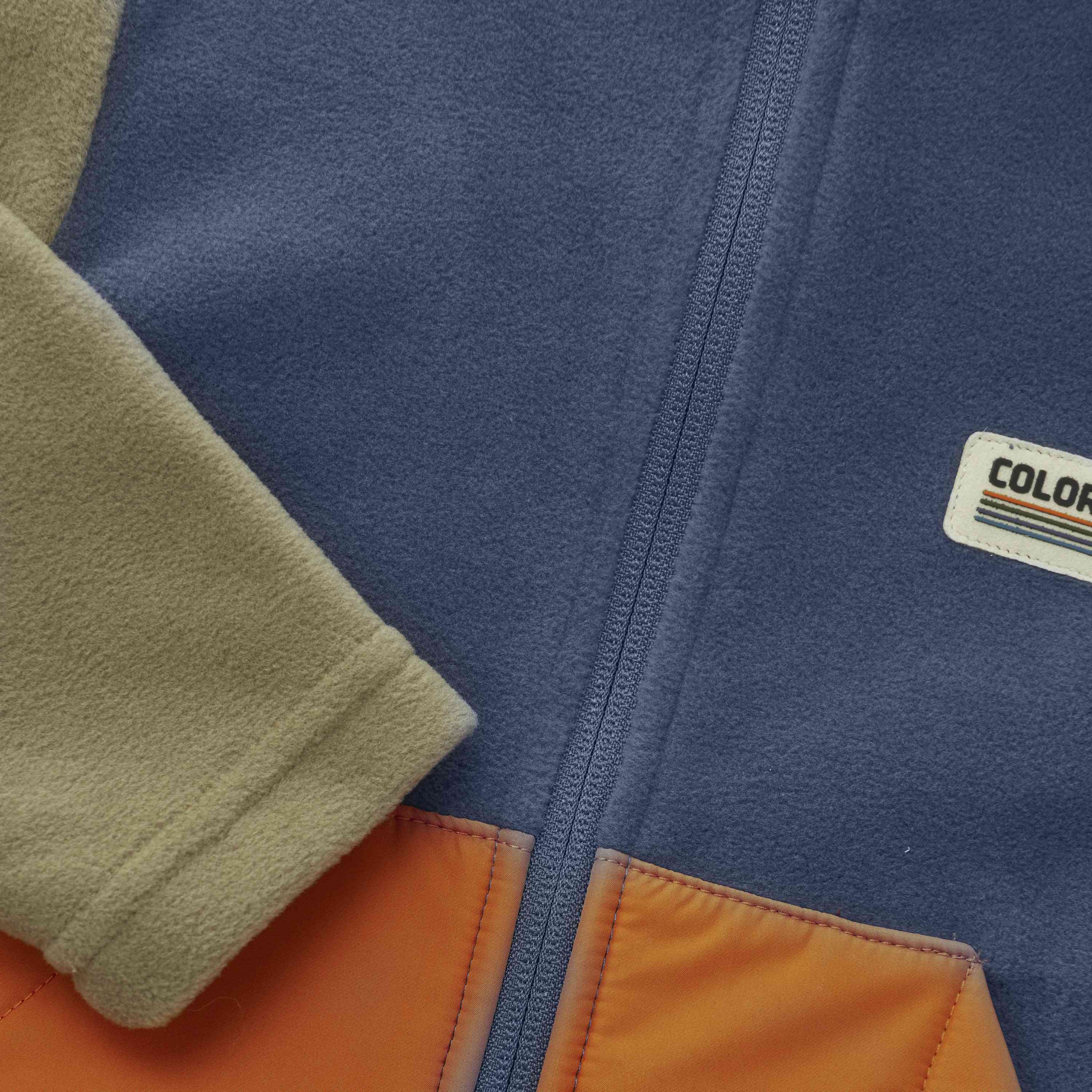 Nahaufnahme einer Color Kids - Baby Fleecejacke mit beigen Ärmeln, blauem Fleecekörper und orangefarbenem Stoff am unteren Ende. Die Jacke hat einen sichtbaren Reissverschluss und einen kleinen rechteckigen Aufnäher mit dem Wort „COLOR“ und einem minimalistischen Streifenmuster auf der Brust. Weitere Details finden Sie in der folgenden Beschreibung.