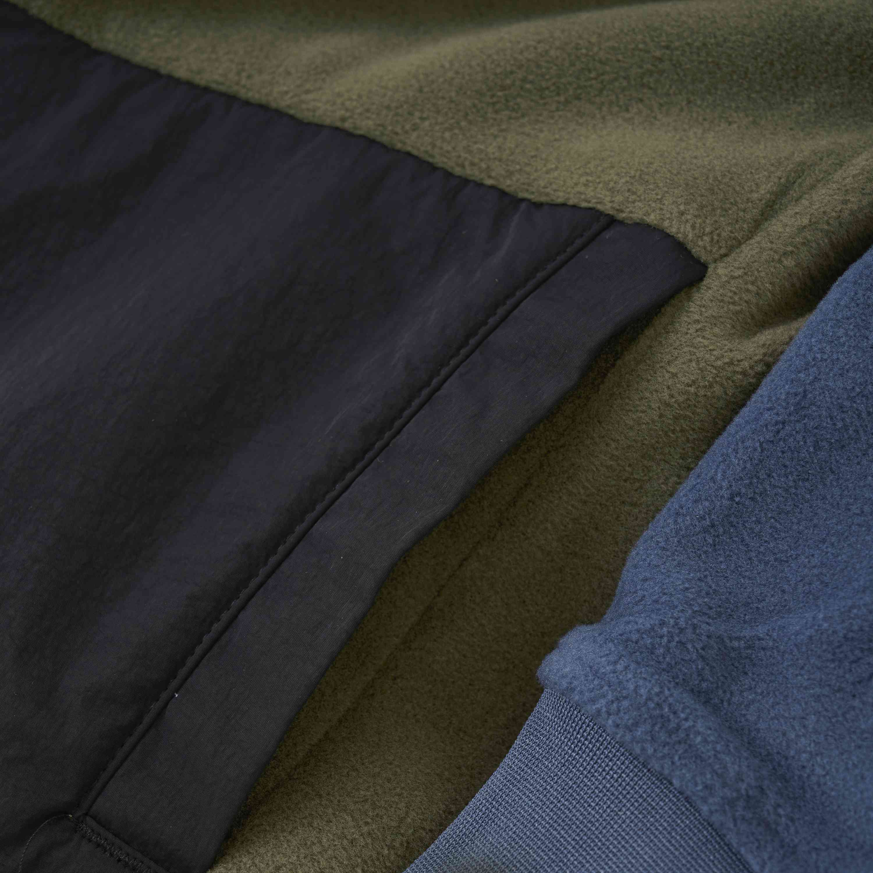 Nahaufnahme der Color Kids - Fleecejacke - Colorblock von Color Kids, die drei verschiedene Stoffe und Farben zeigt. Der linke Abschnitt besteht aus olivgrünem Fleece, der mittlere Abschnitt aus schwarzem Synthetikmaterial mit sichtbaren Nähten und der rechte Abschnitt aus blauem Fleece. Die Texturen sind glatt und gemütlich.