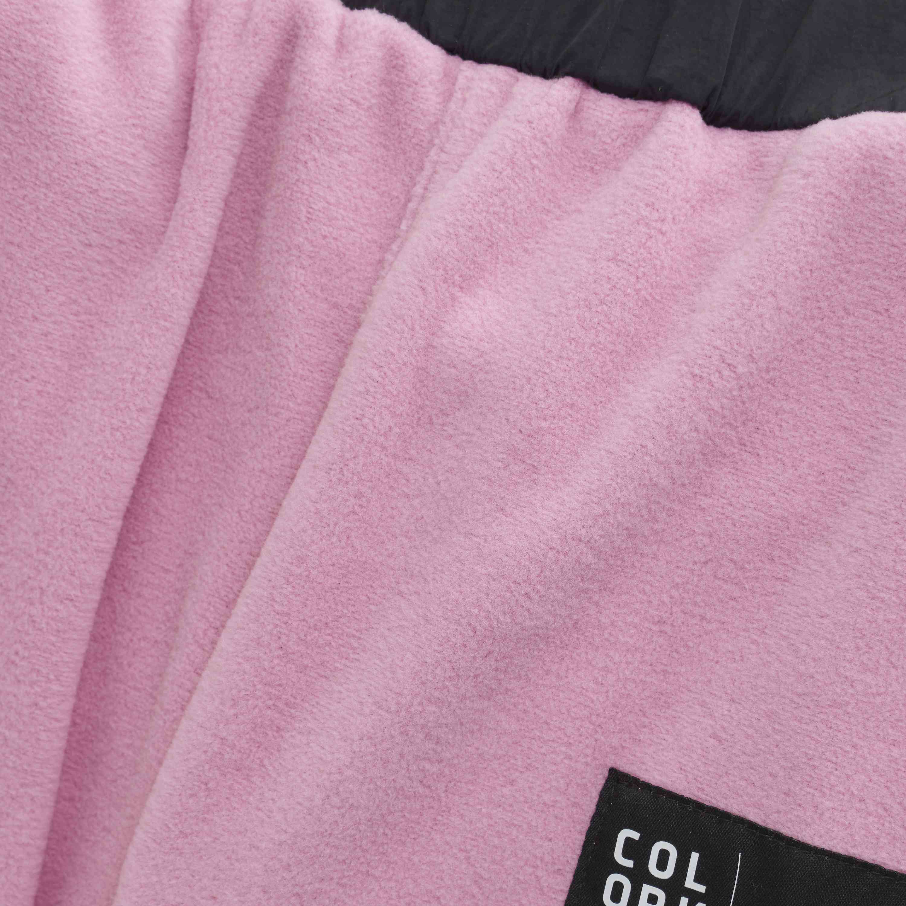 Nahaufnahme eines weichen, rosa Stoffs „Color Kids - Fleece Hosen“, auf den ein Teil eines schwarzen Etiketts aufgenäht ist. Auf dem Etikett ist teilweise weisser Text zu sehen, der die Buchstaben „COL OR“ zeigt. Am oberen Bildrand ist die Oberkante eines dunkleren Stoffs zu sehen.