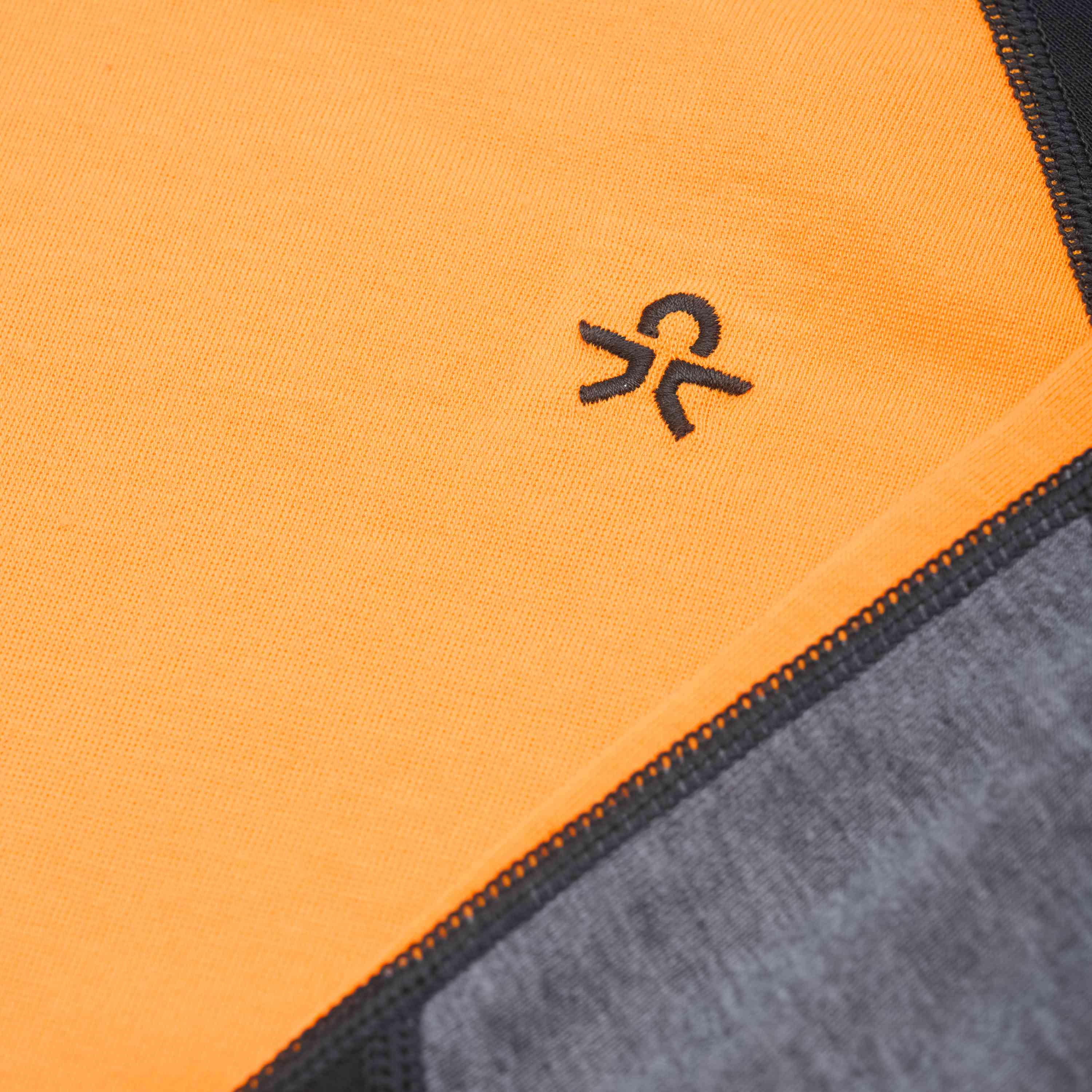 Nahaufnahme des Color Kids - Skiunterwäsche - Colorblock-Kleidungsstücks mit einem orangefarbenen Abschnitt mit einem kleinen, gestickten schwarzen Logo, das einem stilisierten „OK“-Symbol ähnelt. Angrenzende Stoffpaneele in Grau und Schwarz mit auffälligen Nähten. Beschreibung folgt.