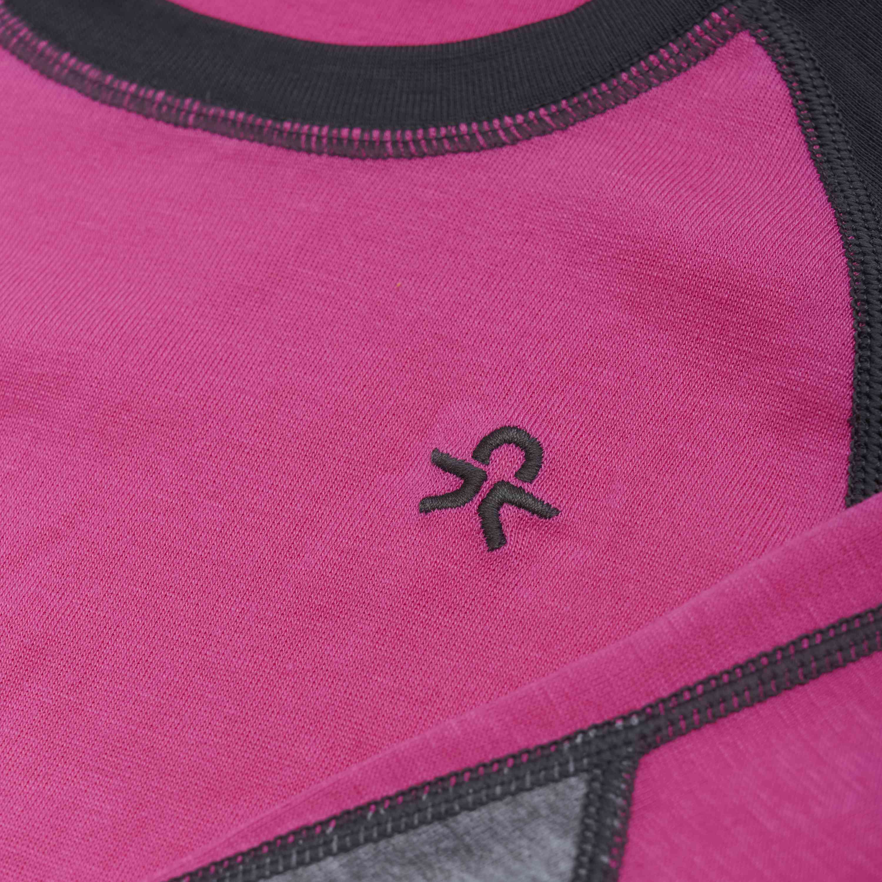 Nahaufnahme eines leuchtend magentafarbenen Color Kids - Skiunterwäsche - Colorblock-Kleidungsstücks mit schwarzem Rand und kubischem Zickzack-Stichdetail. In der Mitte befindet sich ein gesticktes schwarzes Symbol, das einem stilisierten „R“ ähnelt. Der strukturierte und weiche Stoff folgt eng der traditionellen Handwerkskunst von Color Kids.
