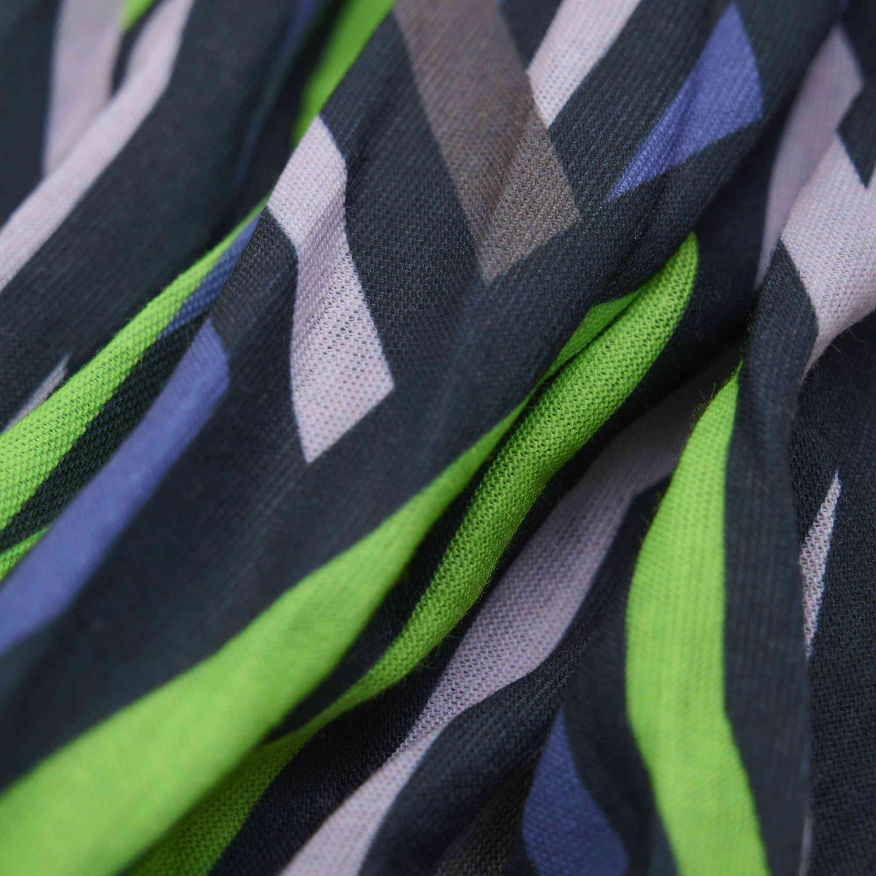 Nahaufnahme des Color Kids Neckwarmer – AOP der Marke Color Kids, der einen Stoff mit einem farbenfrohen geometrischen Muster zeigt. Das Design weist diagonale Streifen in den Farbtönen Grün, Lila, Blau, Schwarz, Grau und Weiss auf. In dieser Beschreibung sieht man einen weichen Stoff mit einer leicht fliessenden und leicht zerknitterten Oberfläche.