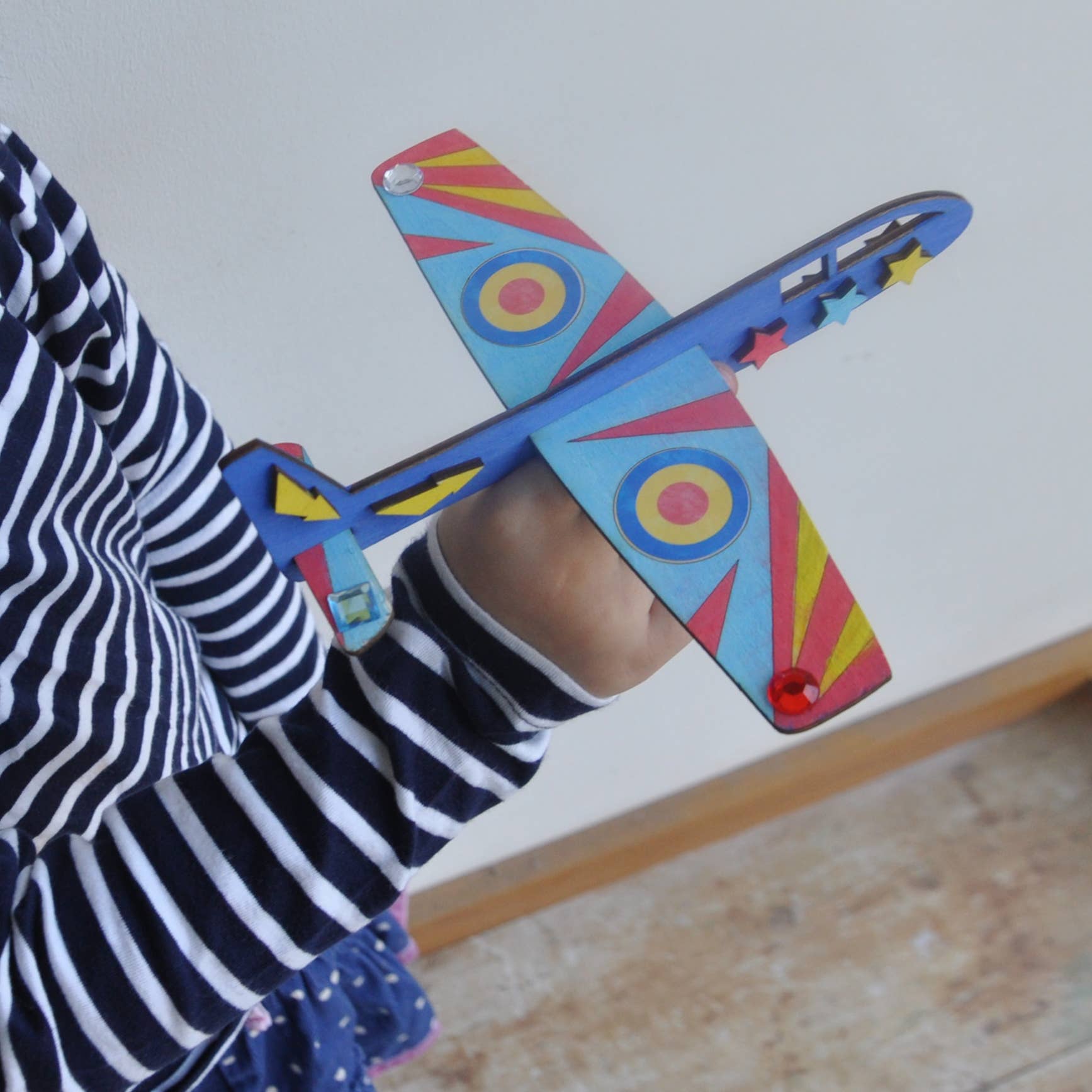 Ein Kind in einem langärmeligen Hemd mit schwarzen und weißen Streifen hält ein buntes Spielzeugflugzeug aus der Cotton Twist - DIY Flugzeug-Set Activity Box. Das Flugzeug ist mit Kreisen, Pfeilen und Sternen in Rot, Blau, Gelb und Rosa gestaltet. Der Hintergrund ist eine schlichte Inneneinrichtung mit Holzboden und weißer Wand.