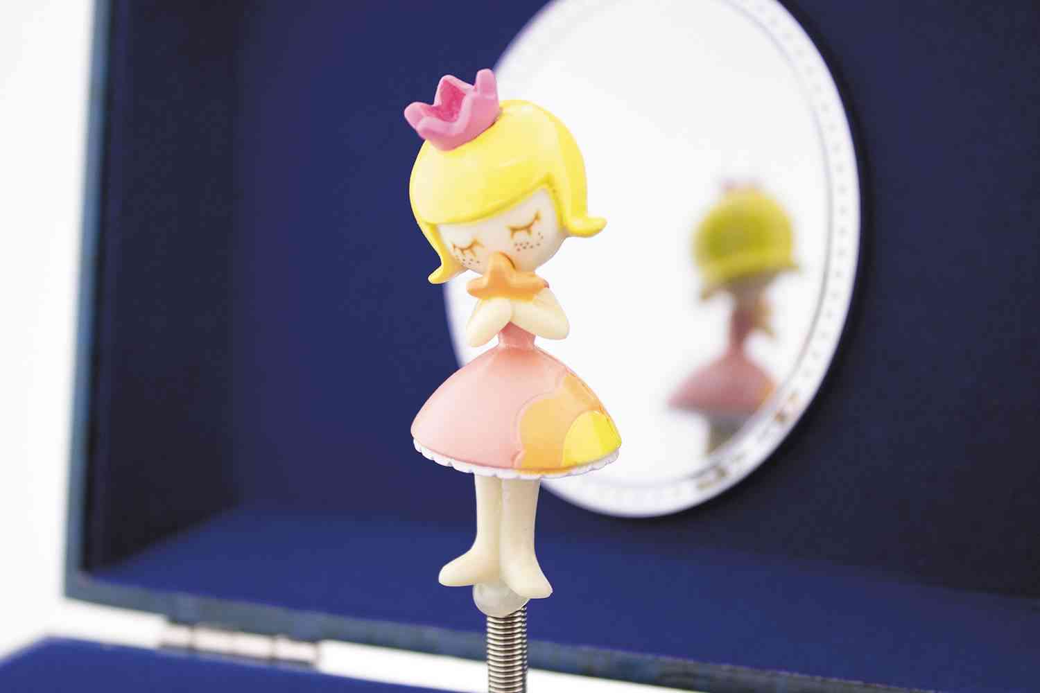 Eine kleine Figur eines blonden Mädchens in einem rosa Kleid mit geschlossenen Augen und vor dem Mund gefalteten Händen steht vor einem runden Spiegel, der ihr Bild reflektiert. Dieses bezaubernde Stück ist ein hervorragendes Beispiel für Ulysse Couleurs d'Enfance - Musiktruhe Frosch von Ulysse Couleurs d'Enfance. Der Hintergrund ist dunkelblau.