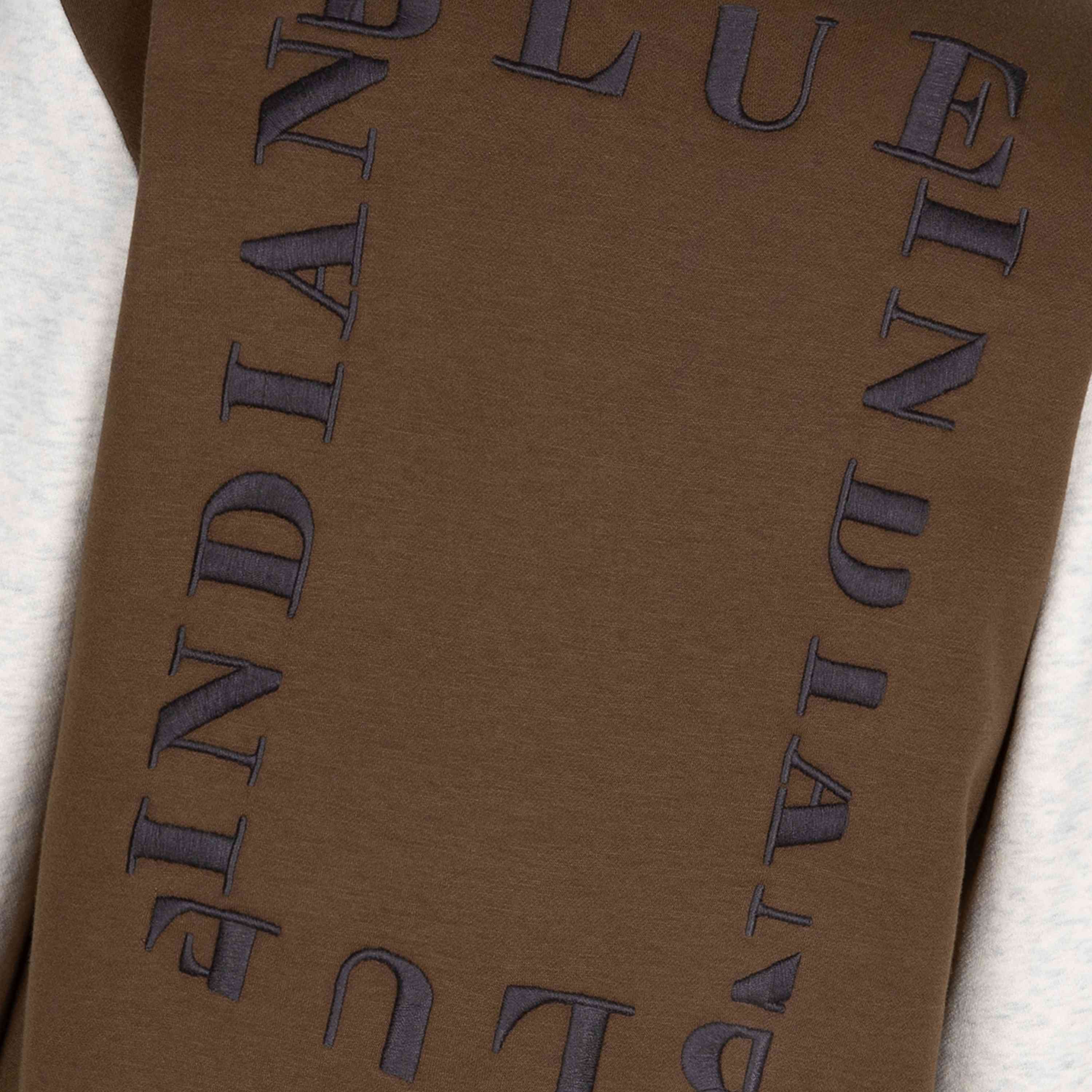 Eine Nahaufnahme eines braunen Stoffes der Indian Blue Jeans - Bomber Jacket Indian, mit dem Text „INDIAN BLUE JEANS“, der in dunkler Stickerei wiederholt um den Umfang gestickt ist und einen rechteckigen Rahmen bildet. Der mittlere Teil des Stoffes ist hellgrau. Beschreibung folgt.