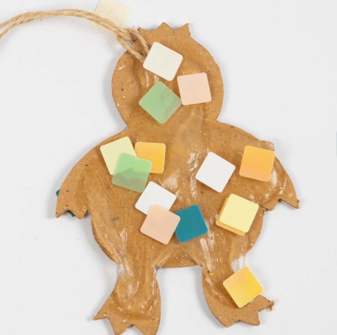 Ein Bastelprojekt in Form eines Kükens aus brauner Pappe ist mit kleinen, bunten quadratischen Papierstücken verziert. Am oberen Ende des Kükens ist eine Schnur zum Aufhängen befestigt. Mit diesem Creativ Company - Oster-DIY-Set für Kinder - Mosaik können sich Kinder ganz einfach kreativ betätigen. Der Hintergrund ist schlicht weiß.