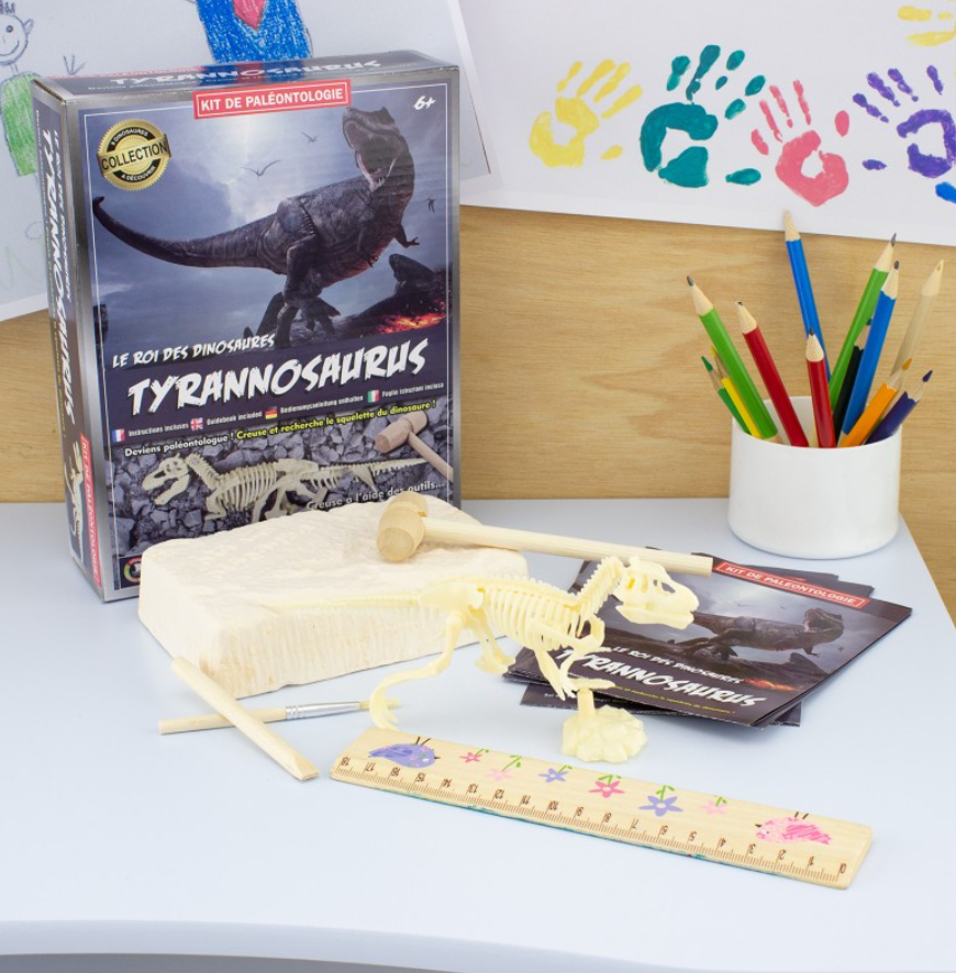 Das Ulysse Couleurs d'Enfance - Entdeckungsspielzeug: Dinosaurier von Ulysse Couleurs d'Enfance mit einem Tyrannosaurus-Skelettmodell wird ausgestellt. Das Set enthält eine Schachtel, Skelettbauteile, ein Lineal mit farbenfrohen Dinosaurierillustrationen, Ausgrabungswerkzeuge zum Freilegen vergrabener Skelette, ein Heft und Buntstifte in einem weißen Becher. Zeichnungen hängen an der Wand.