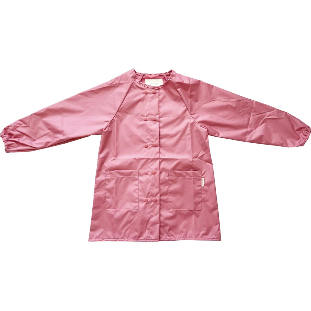 Auf weißem Hintergrund ist ein langärmliger, rosa Grech & Co. - Malkittel für Kinder aus recyceltem Stoff abgebildet. Der Kittel hat ein schlichtes Design mit elastischen Bündchen, zwei großen Vordertaschen und einem kleinen Hängeetikett an einer Seite.