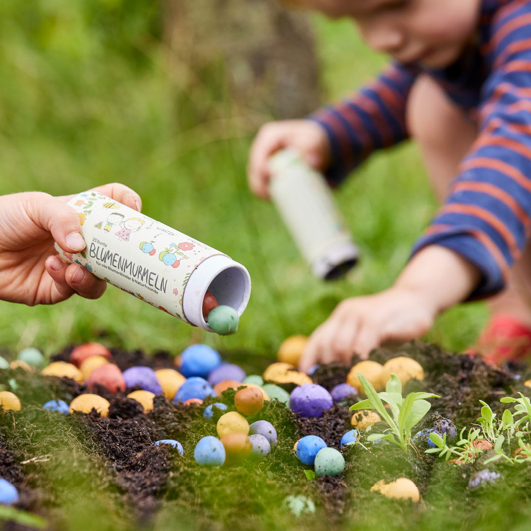 Eine Nahaufnahme eines Kindes, das bunte Blumenmurmeln in Dose von Die Stadtgärnter aus einem Behälter auf die Erde in einem Garten schüttet. Ein anderes Kind, im Hintergrund leicht verschwommen, pflanzt ebenfalls Samenkugeln. Verschiedene farbenfrohe Samenkugeln sind auf dem Boden zwischen Gras und kleinen Pflanzen verstreut, eine perfekte Szene für Die Stadtgärnter Nordhorn.